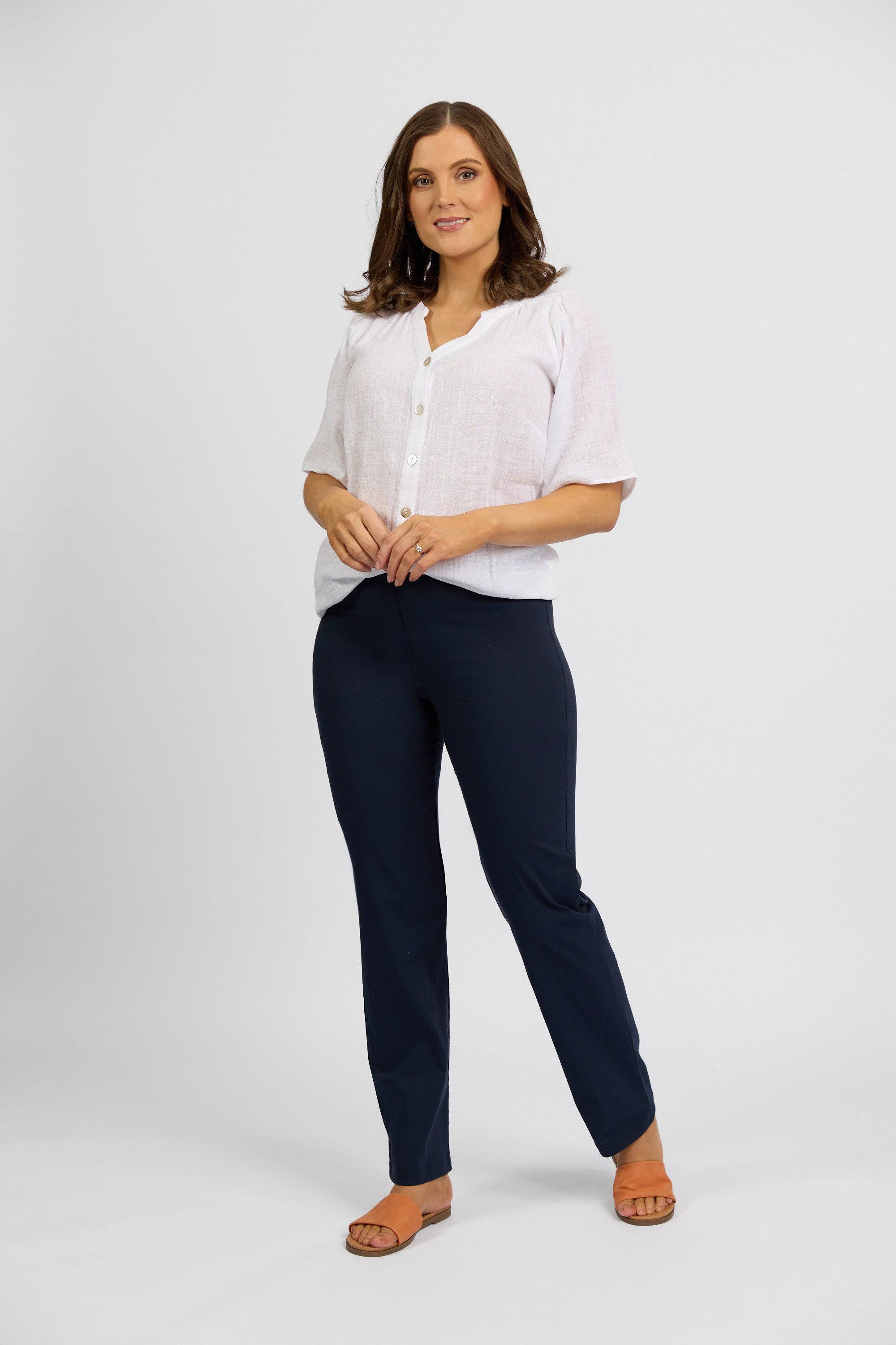 A woman with shoulder-length brown hair stands against a white background, smiling slightly with her hands clasped. She wears a white blouse and Vassalli Paris Full Length Pant - Inka in navy blue.