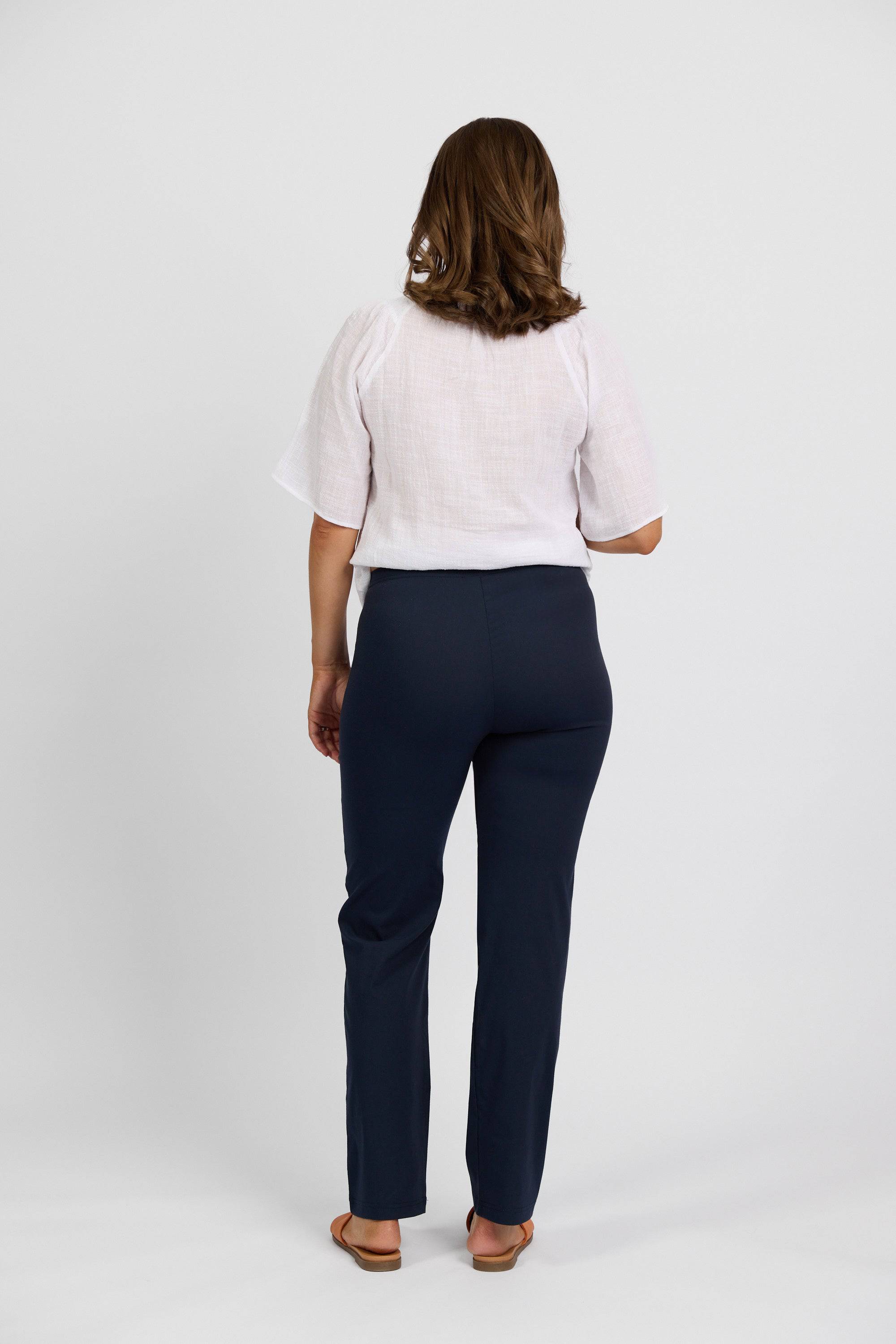 A woman faces away from the camera wearing a white blouse, sandals, and Vassalli Paris Full Length Pant - Inka in navy stretch fabric, against a plain white background.