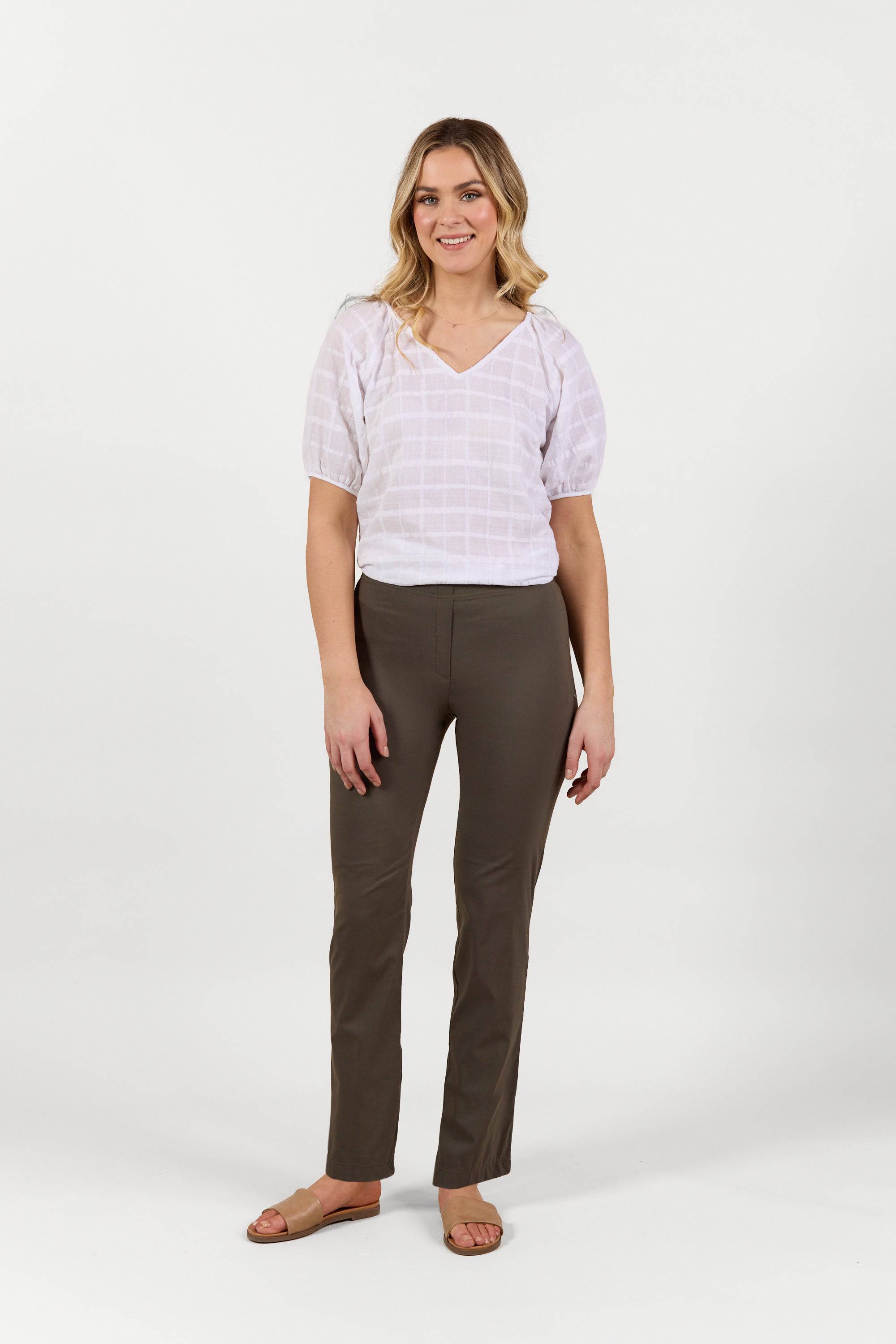 A woman stands against a white background, smiling in a white short-sleeve blouse, Vassalli Paris Full Length Pant - Khaki, and tan sandals. Her loose, wavy hair frames her relaxed pose with arms at her sides.