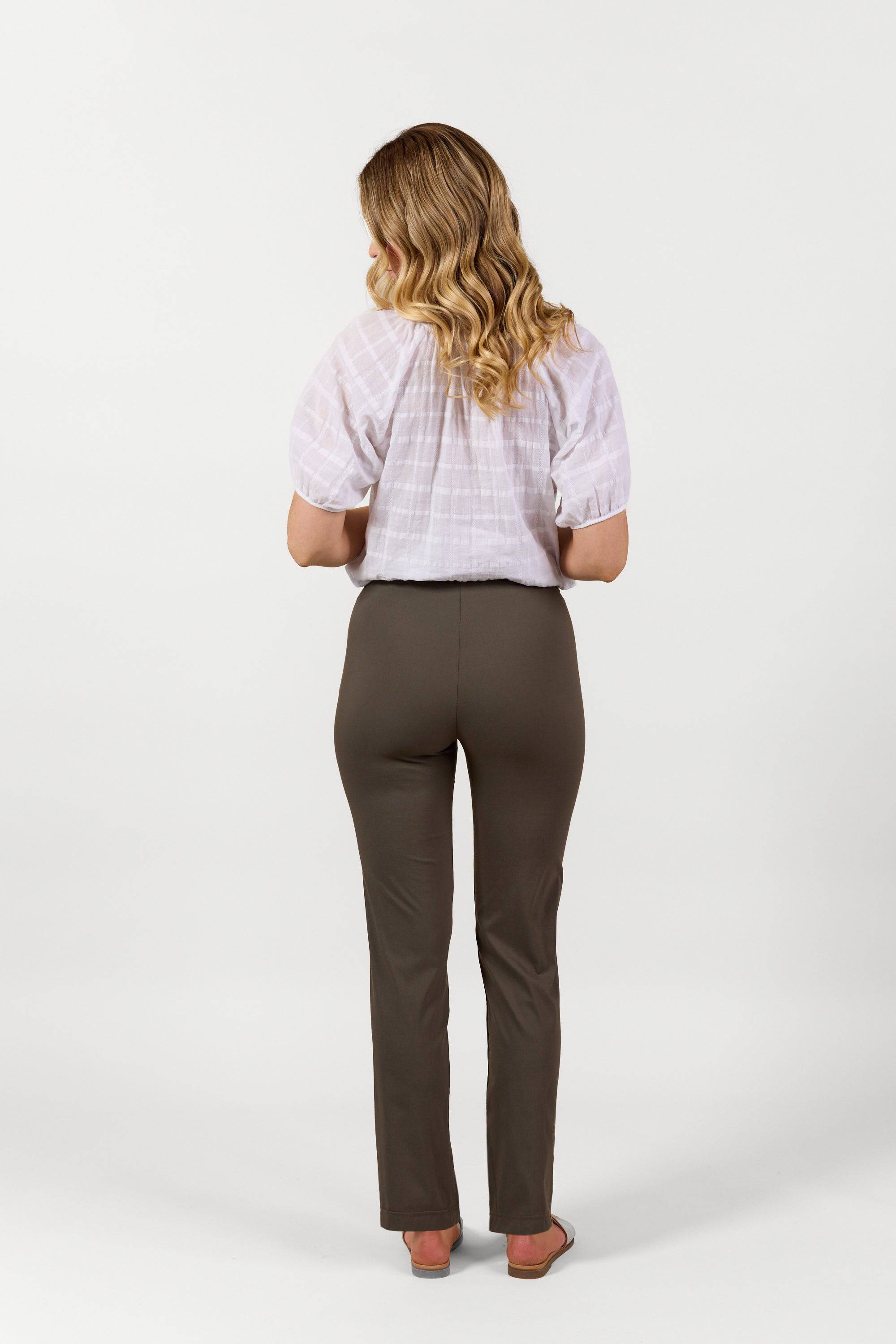 A woman with wavy blonde hair stands facing away, wearing a white short-sleeve blouse, Vassalli Paris Full Length Pant in Khaki, and sandals, against a plain white background.
