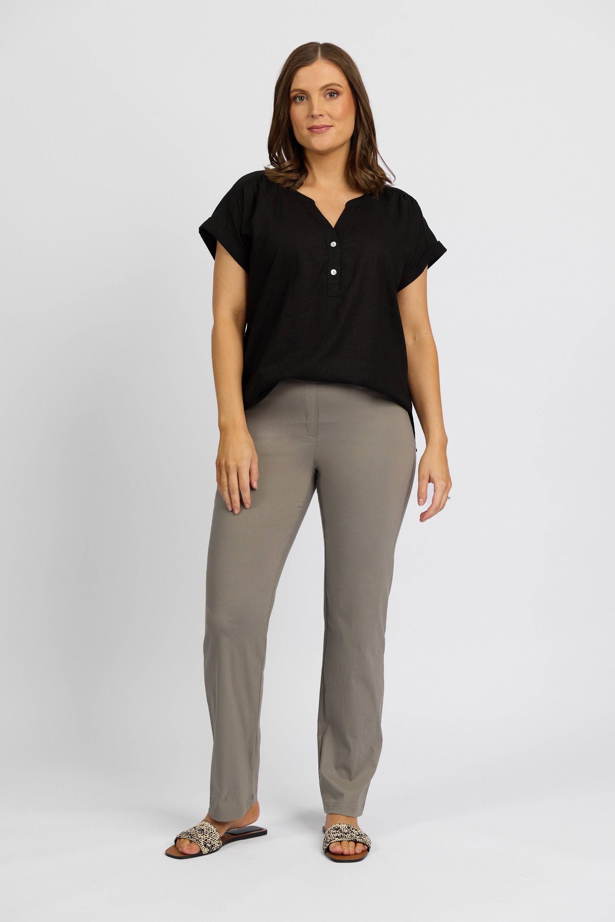 A woman with shoulder-length brown hair and a neutral expression stands against a white background, wearing a black short-sleeve blouse, Vassalli Paris Full Length Pant in Latte, and leopard-print sandals.