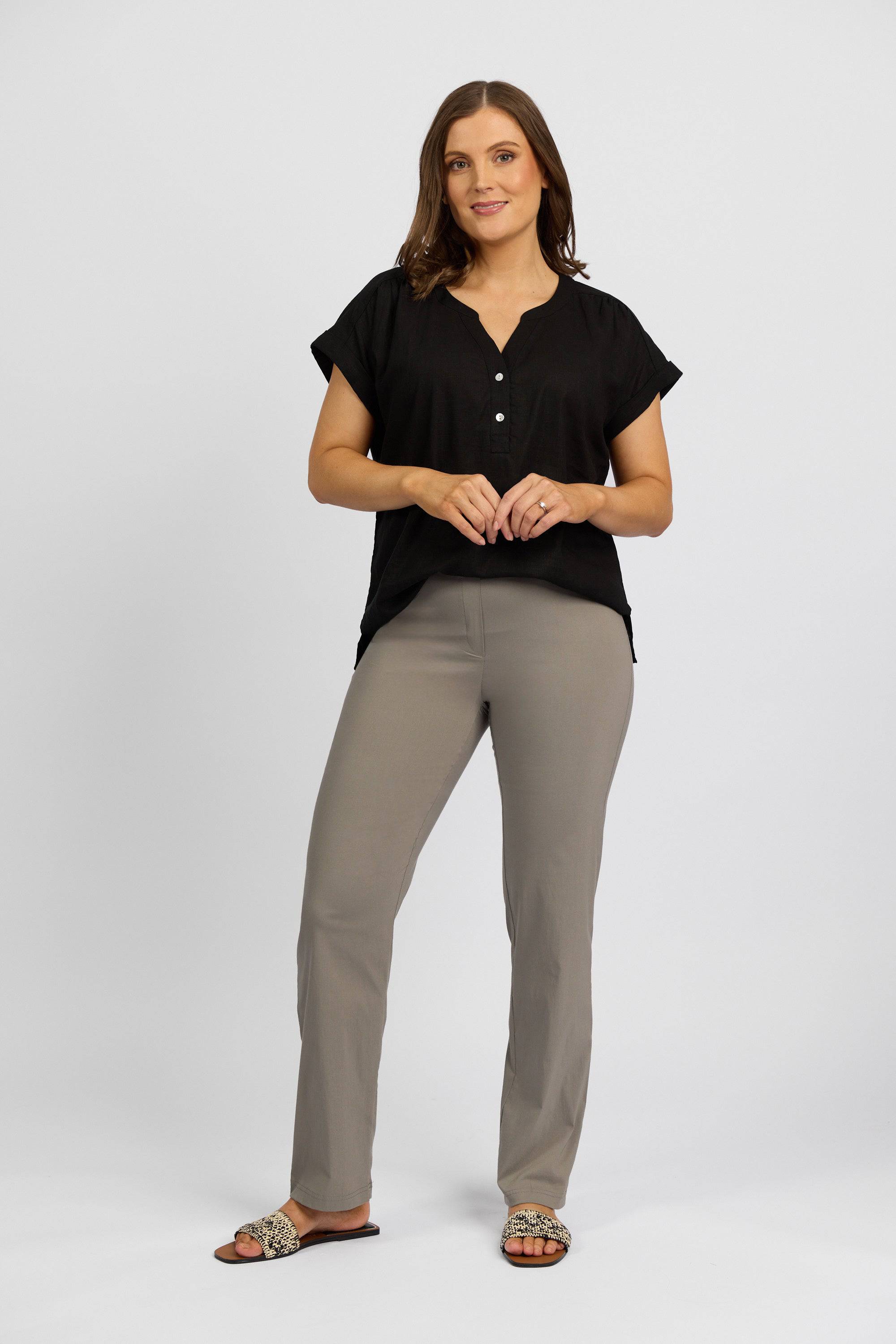 A woman with shoulder-length brown hair stands against a white background, smiling softly in a short-sleeved black button-up top and Vassalli Paris Full Length Pant in Latte. She has her hands together and wears leopard-print slide sandals.