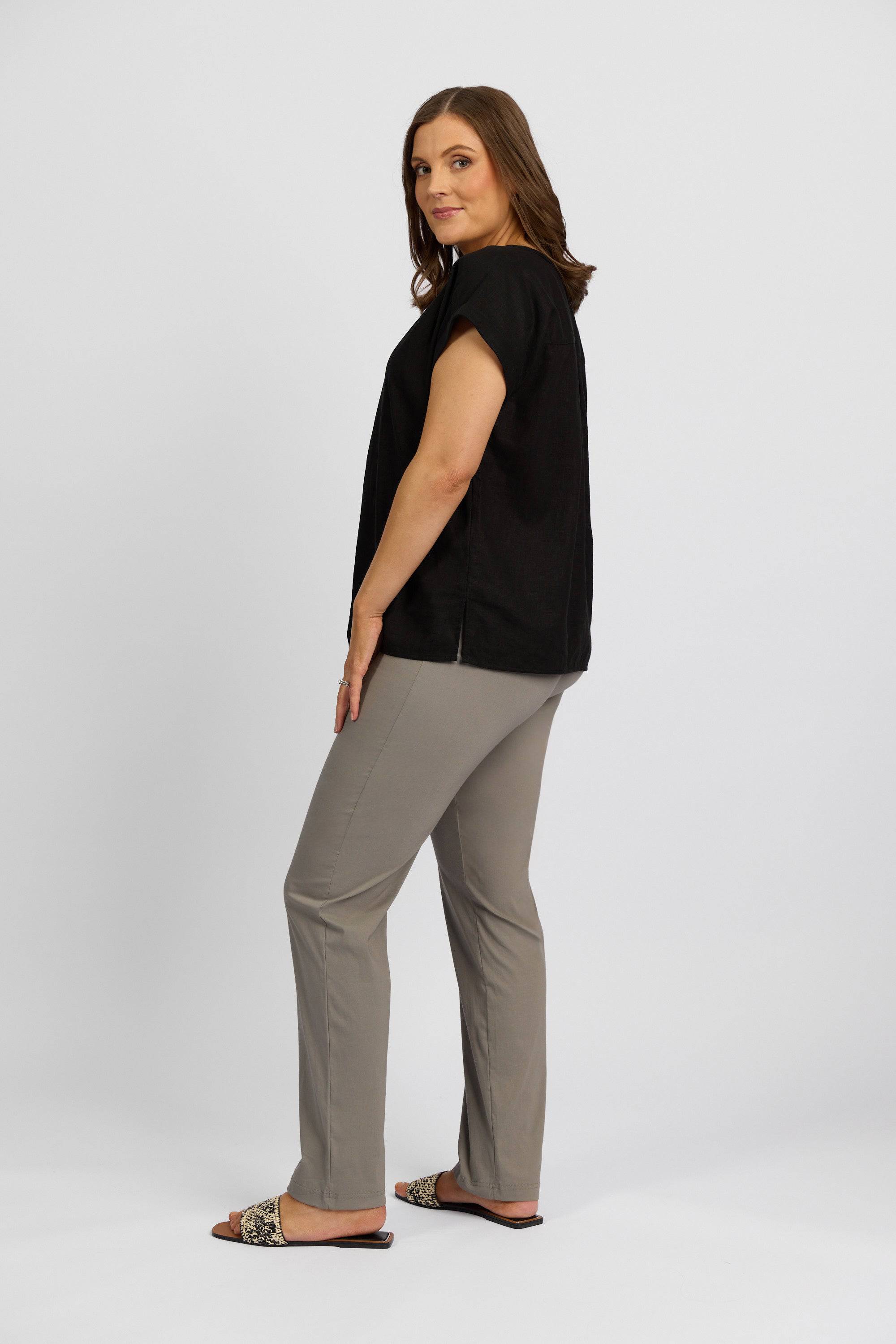 A woman stands in a side pose, wearing a black short-sleeve top, Vassalli Paris Full Length Pant - Latte, and animal-print slide sandals, against a plain white background.