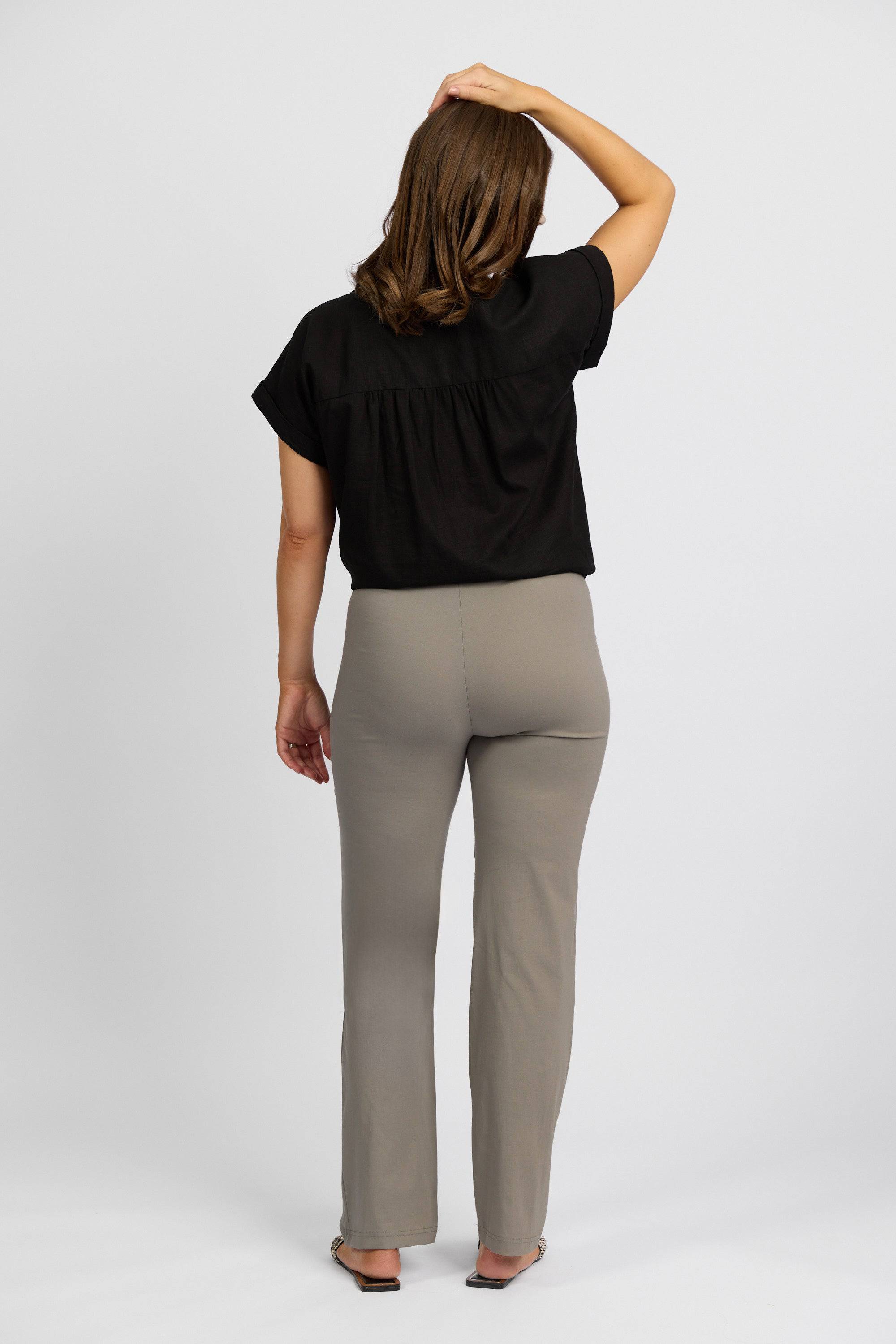 A woman faces away with one arm touching her head, wearing a black short-sleeve top, Vassalli Paris Full Length Pant in Latte, and open-toed shoes against a plain white background.