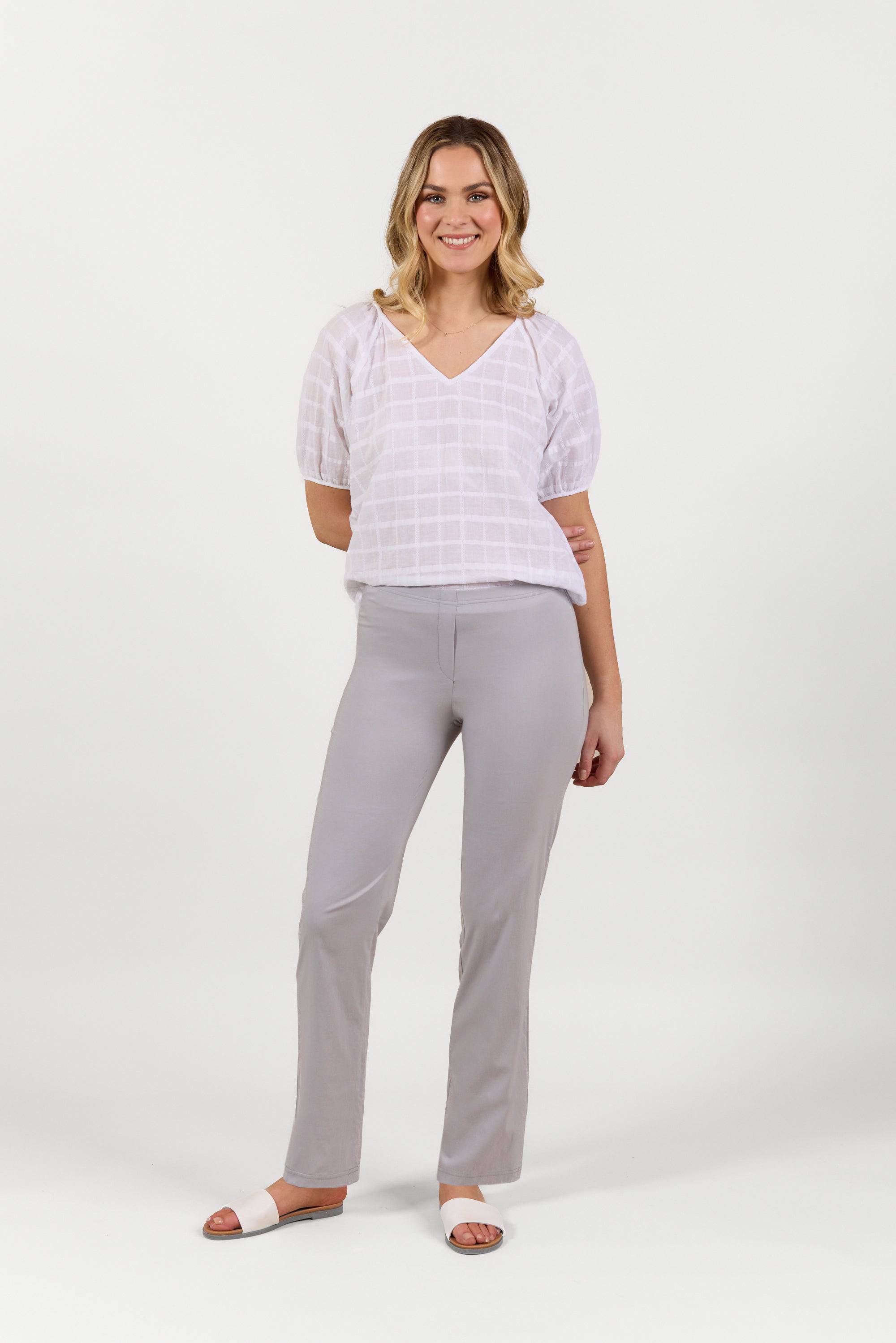 A woman smiles, wearing a white, short-sleeved, V-neck grid blouse, Vassalli Paris Full Length Pant in Silver, and white sandals against a plain white background.