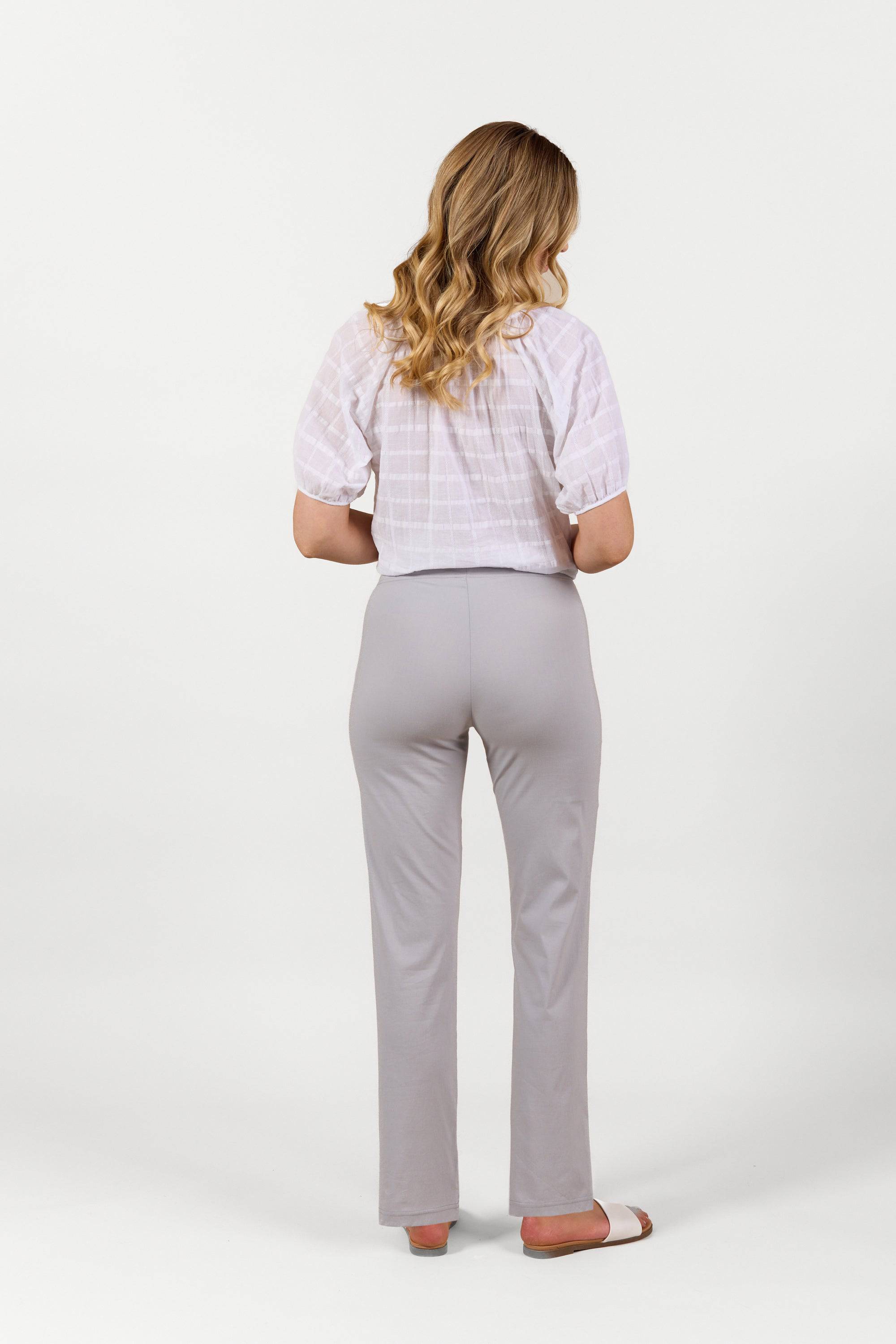 A woman with wavy blonde hair stands facing away, wearing a white short-sleeve blouse, Vassalli Paris Full Length Pant in Silver, and sandals against a plain white background.