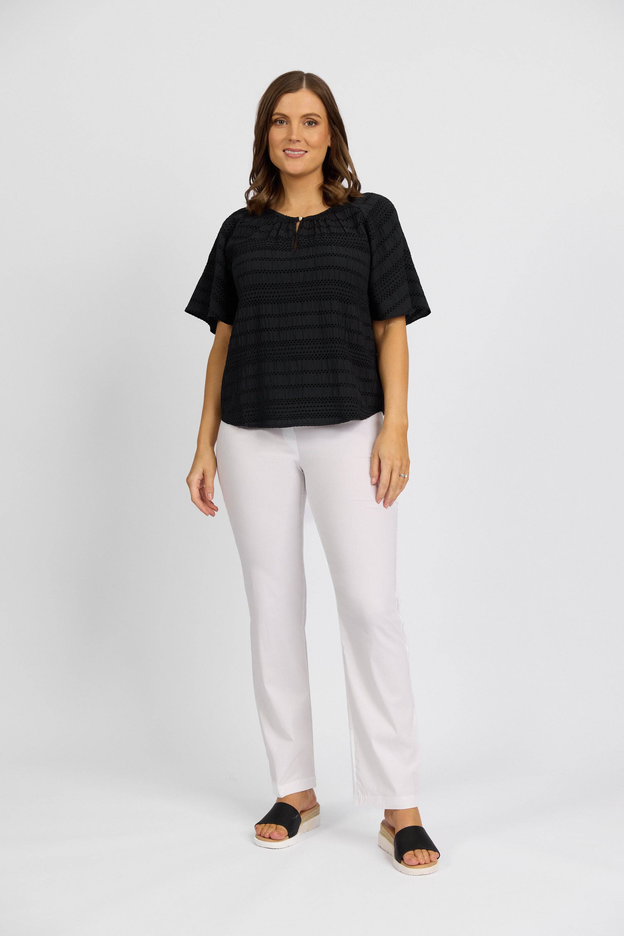 A woman with shoulder-length brown hair smiles against a white background, wearing a loose black short-sleeve top, Vassalli Paris Full Length Pant in white, and black sandals.