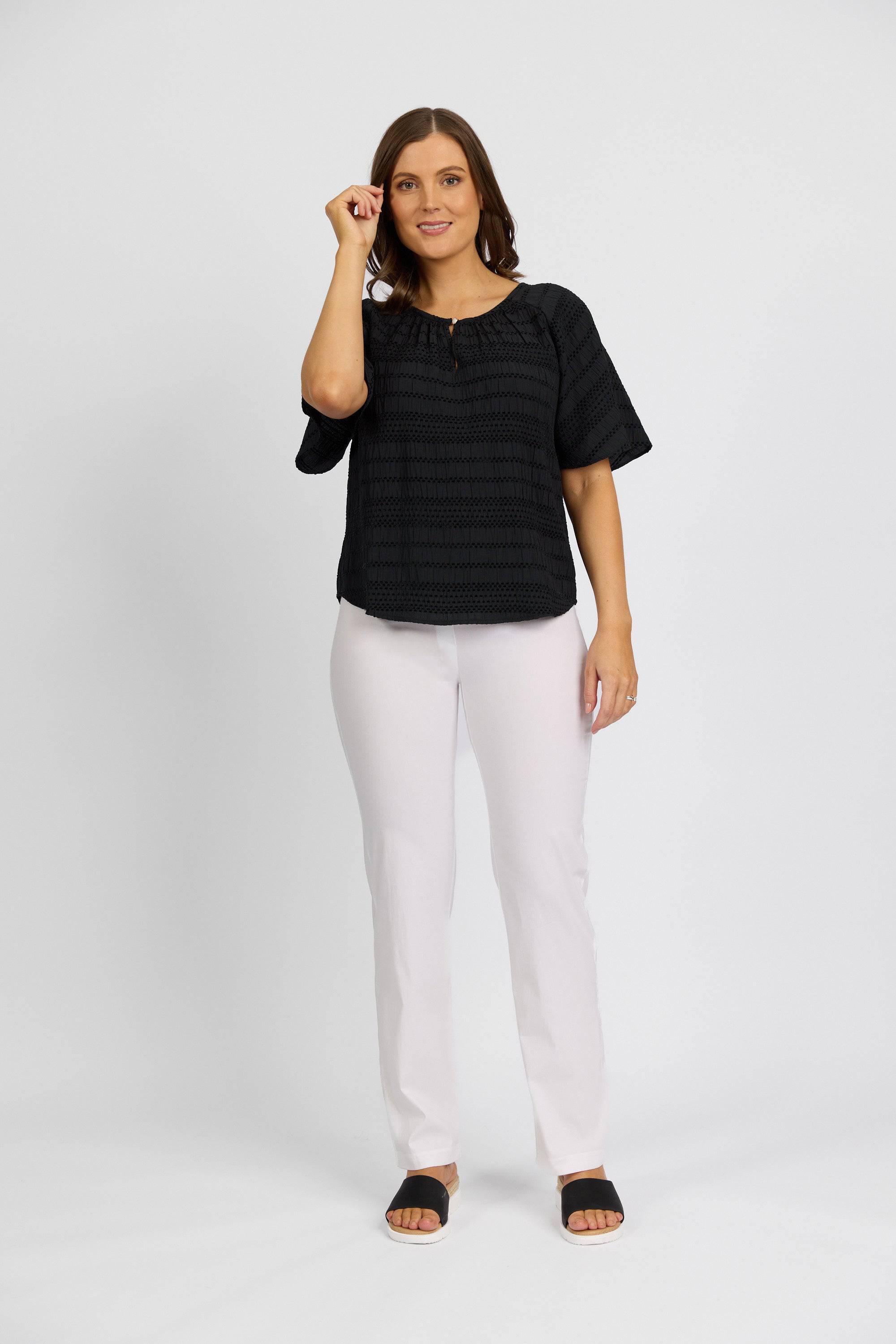 A woman stands against a plain white background, smiling softly with one hand raised, wearing a black short-sleeve blouse, black sandals, and Vassalli's Paris Full Length Pant in white.