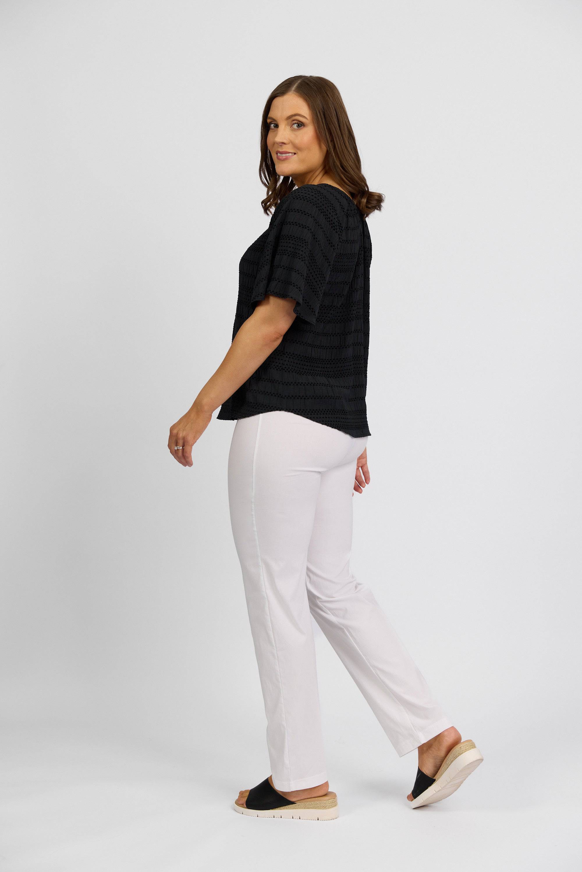 A woman with shoulder-length brown hair, wearing a loose black top, Vassalli Paris Full Length Pant in white, and black slide sandals stands against a plain white background, smiling while looking over her shoulder.