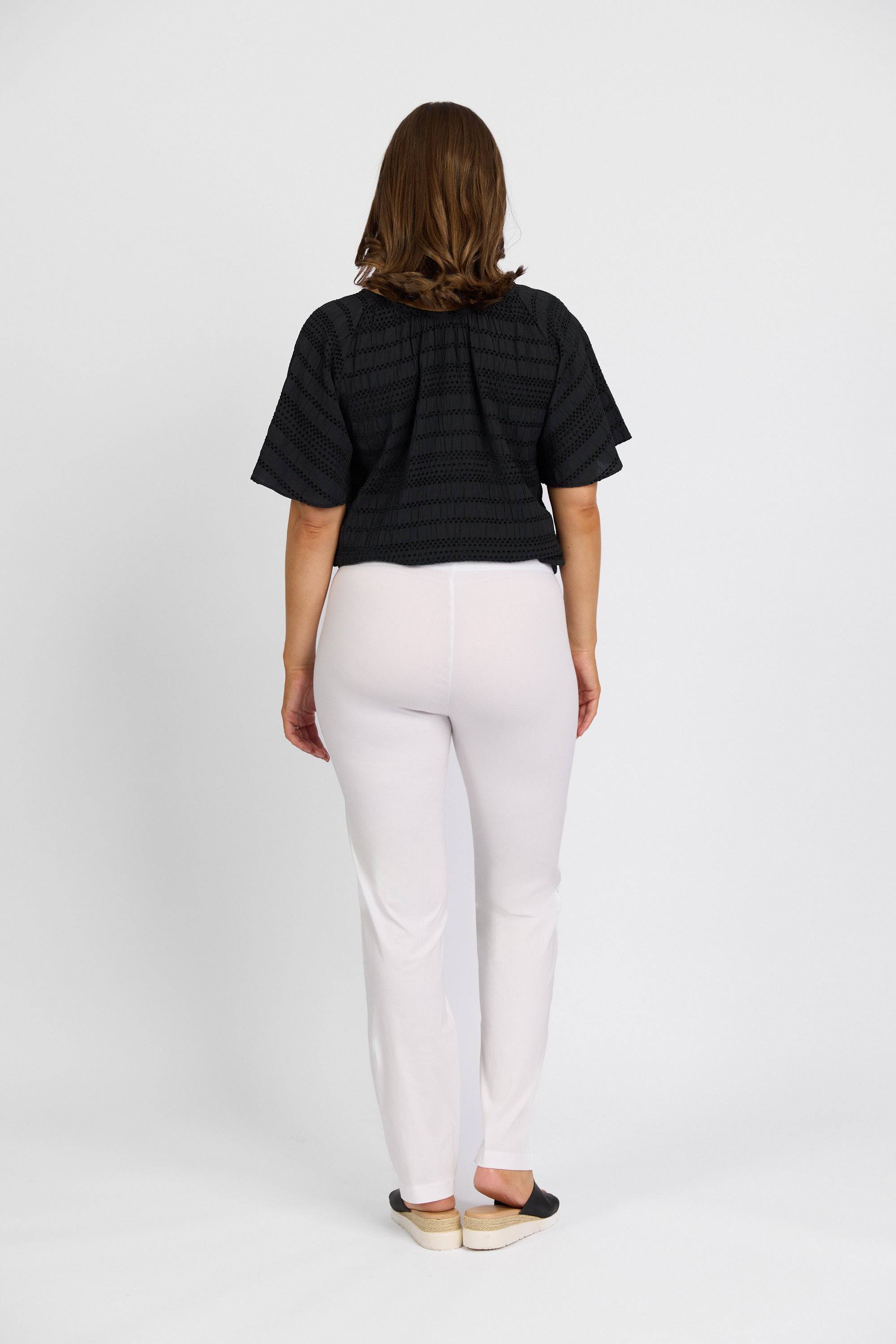 A person with shoulder-length brown hair stands facing away, wearing a black short-sleeve top, Vassalli Paris Full Length Pant in white, and black slip-on shoes against a plain white background.