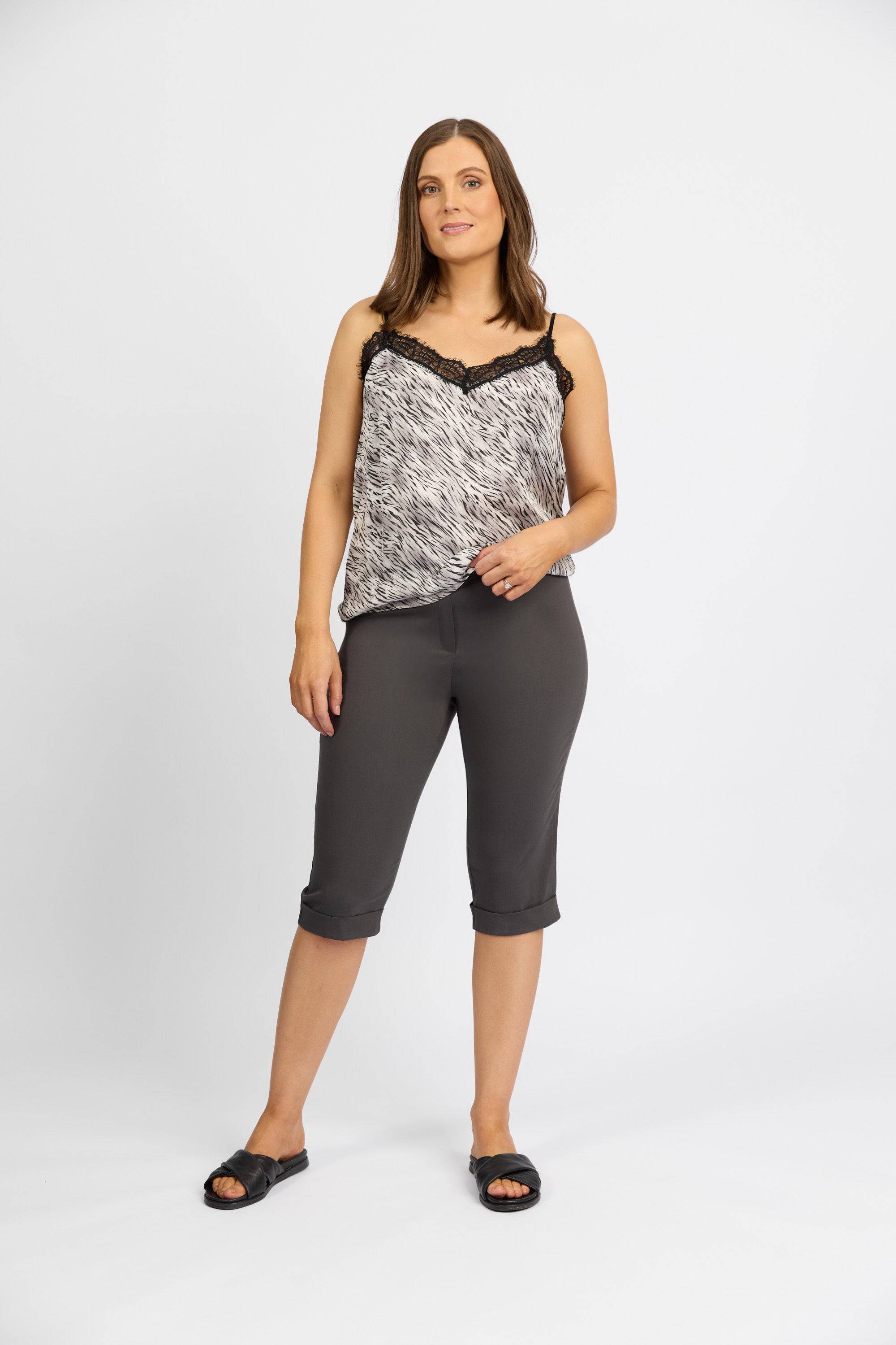 A woman with shoulder-length brown hair and a relaxed expression stands against a white background, wearing Vassalli's Paris Short - Beluga, a black and white sleeveless lace-trimmed top, and black sandals.