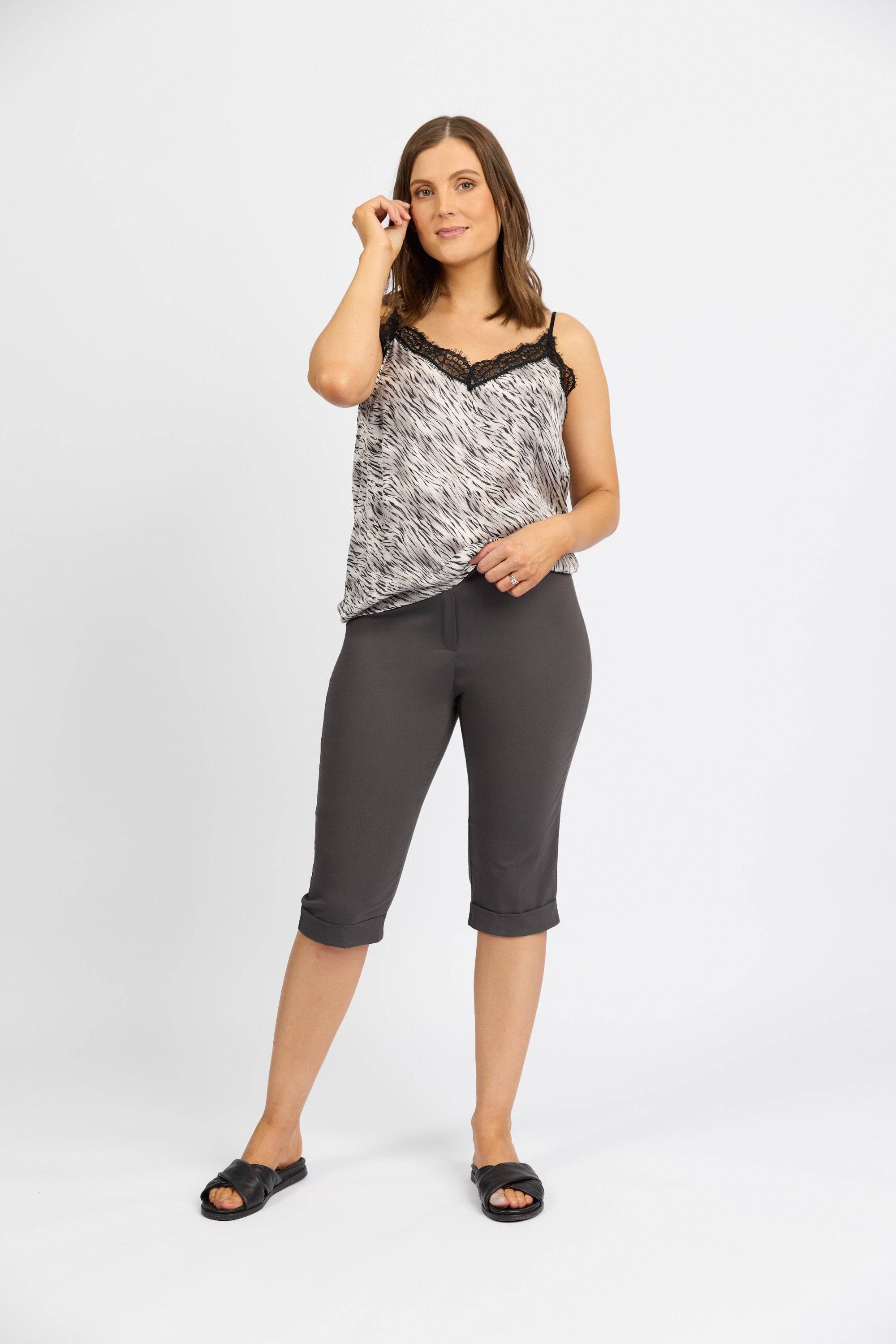 A woman stands against a plain white background, wearing a patterned sleeveless top with lace trim and Vassalli Paris Short - Beluga. She touches her hair and looks at the camera with a slight smile.