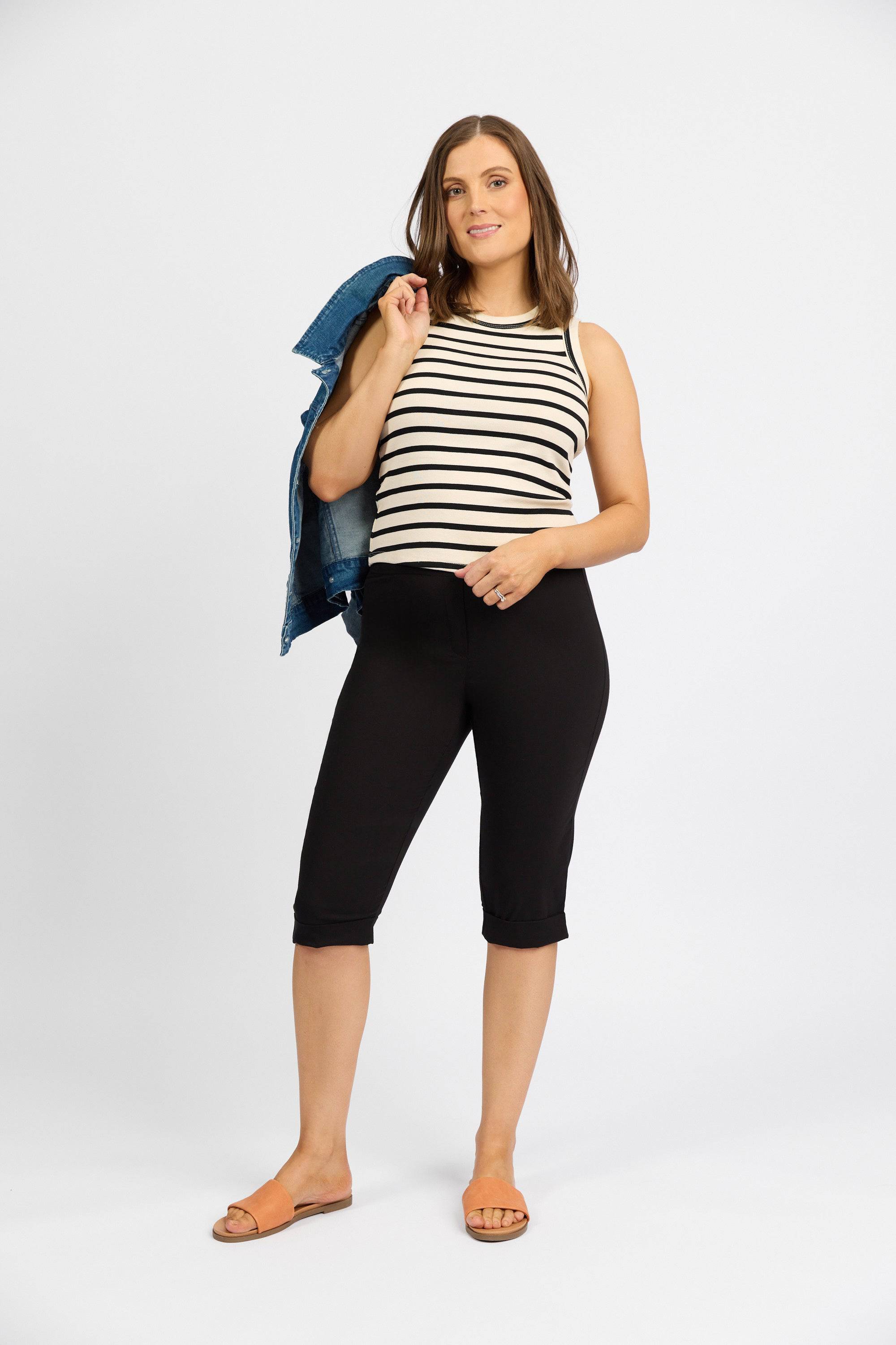 A woman smiles at the camera against a white background, wearing Vassalli’s Paris Short in black, a black and white striped sleeveless stretch top, tan sandals, and holding a denim jacket over her shoulder.