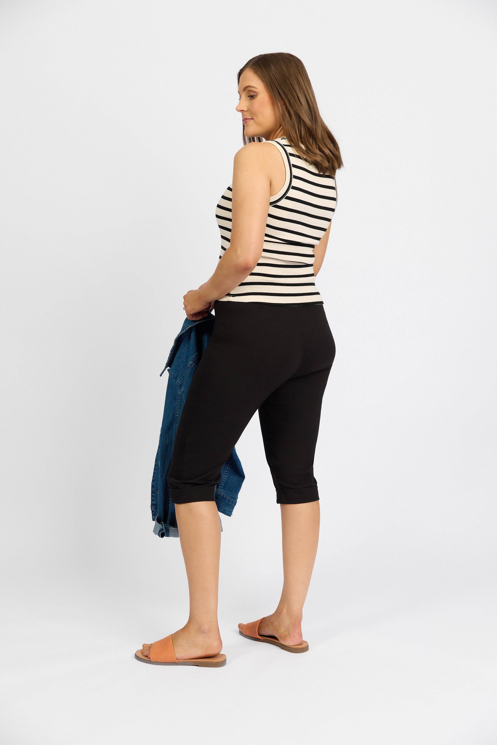 A woman stands sideways against a plain white background, wearing Vassalli's black Paris Shorts with a black-and-white striped stretch tank top, brown sandals, and holding a blue denim jacket.