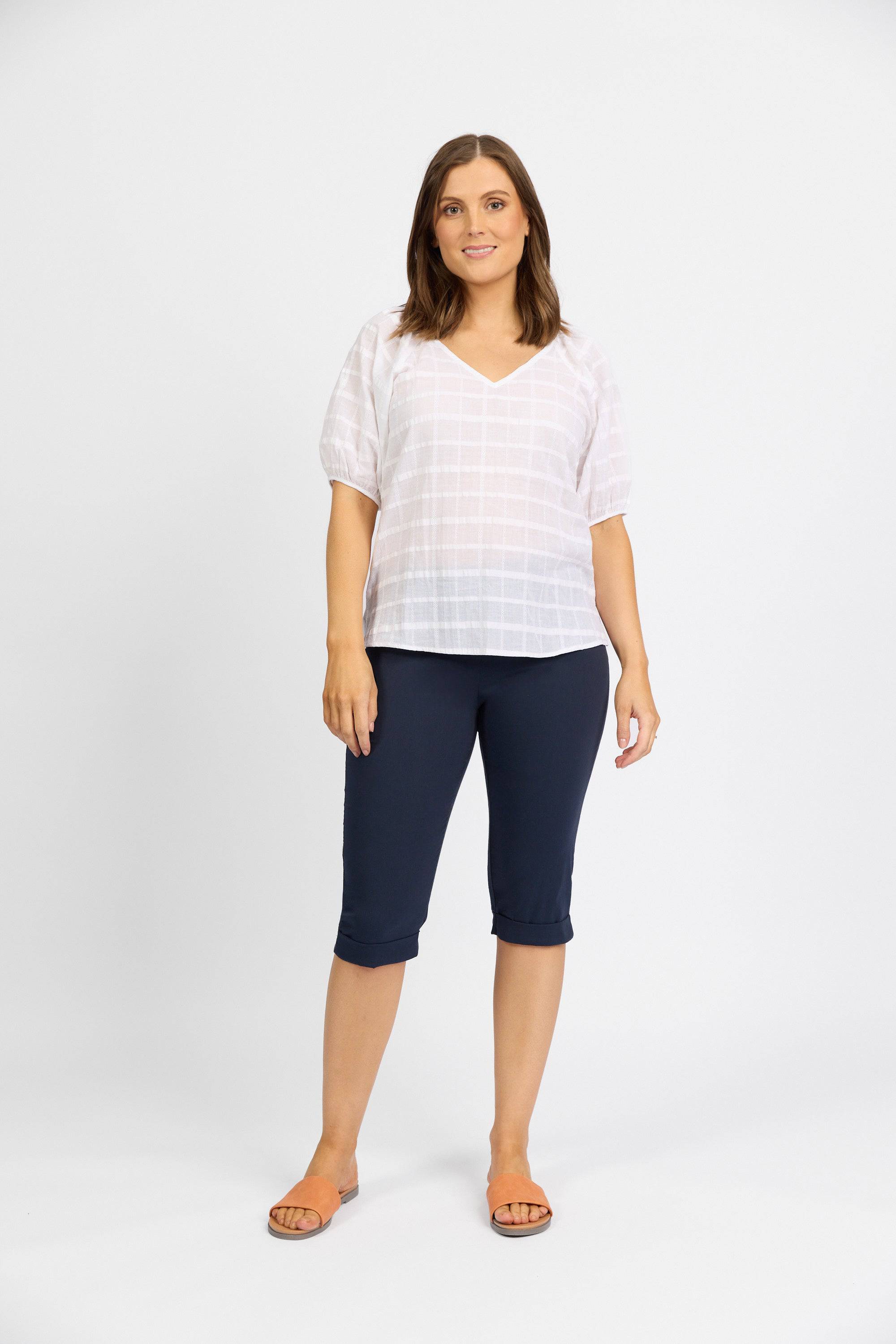 A woman with shoulder-length brown hair stands against a white background, wearing Vassalli's Paris Short - Inka from the Paris Collection, paired with a white short-sleeve blouse and tan sandals. She has a relaxed, neutral expression.