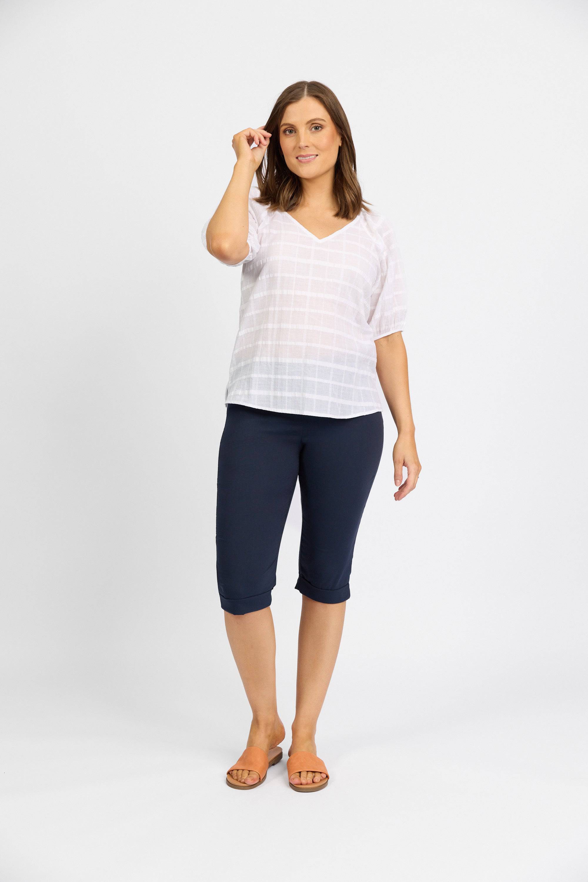 A woman stands against a white background, smiling gently and holding one hand near her face. She wears the Vassalli Paris Short – Inka: navy blue, mid rise, knee-length stretch pants paired with a white short-sleeved top.