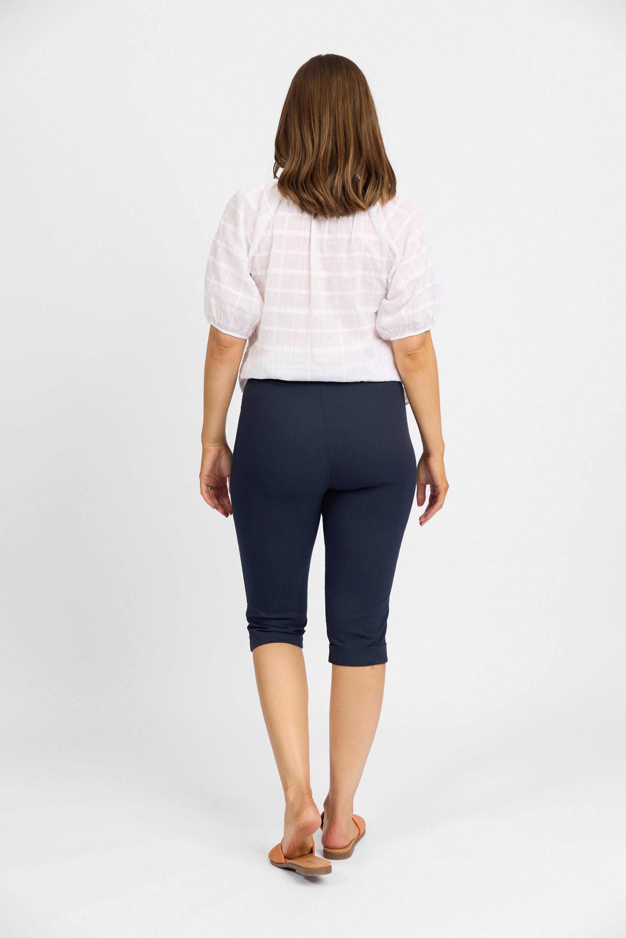 A woman with shoulder-length brown hair, wearing a white short-sleeve blouse, Vassalli Paris Short - Inka, and brown sandals stands facing away on a plain white background.