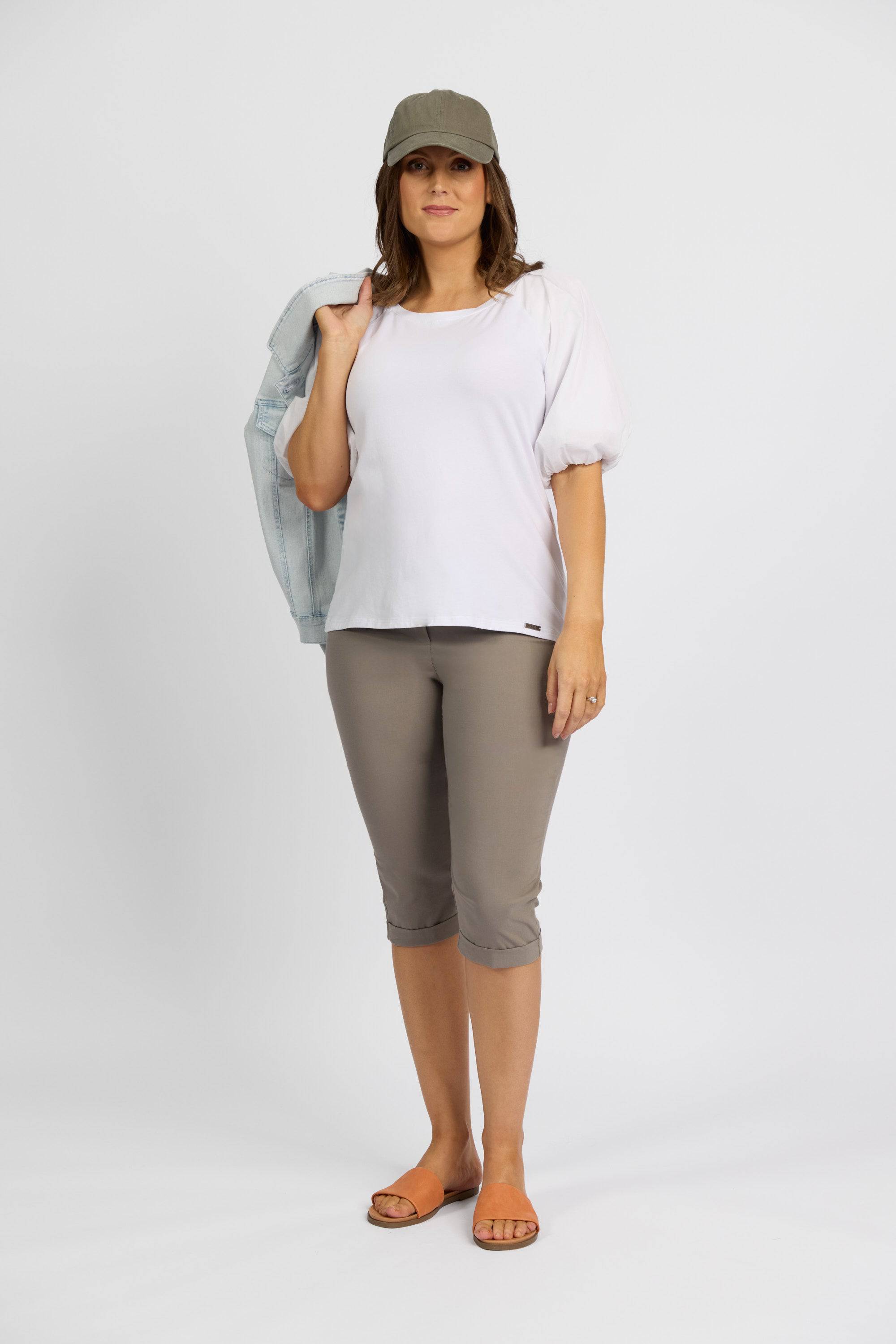 A woman poses against a white backdrop wearing Vassalli’s Paris Short - Latte, a white puff-sleeve top, gray cap, and brown sandals. She holds a light blue jacket over her shoulder and looks at the camera.