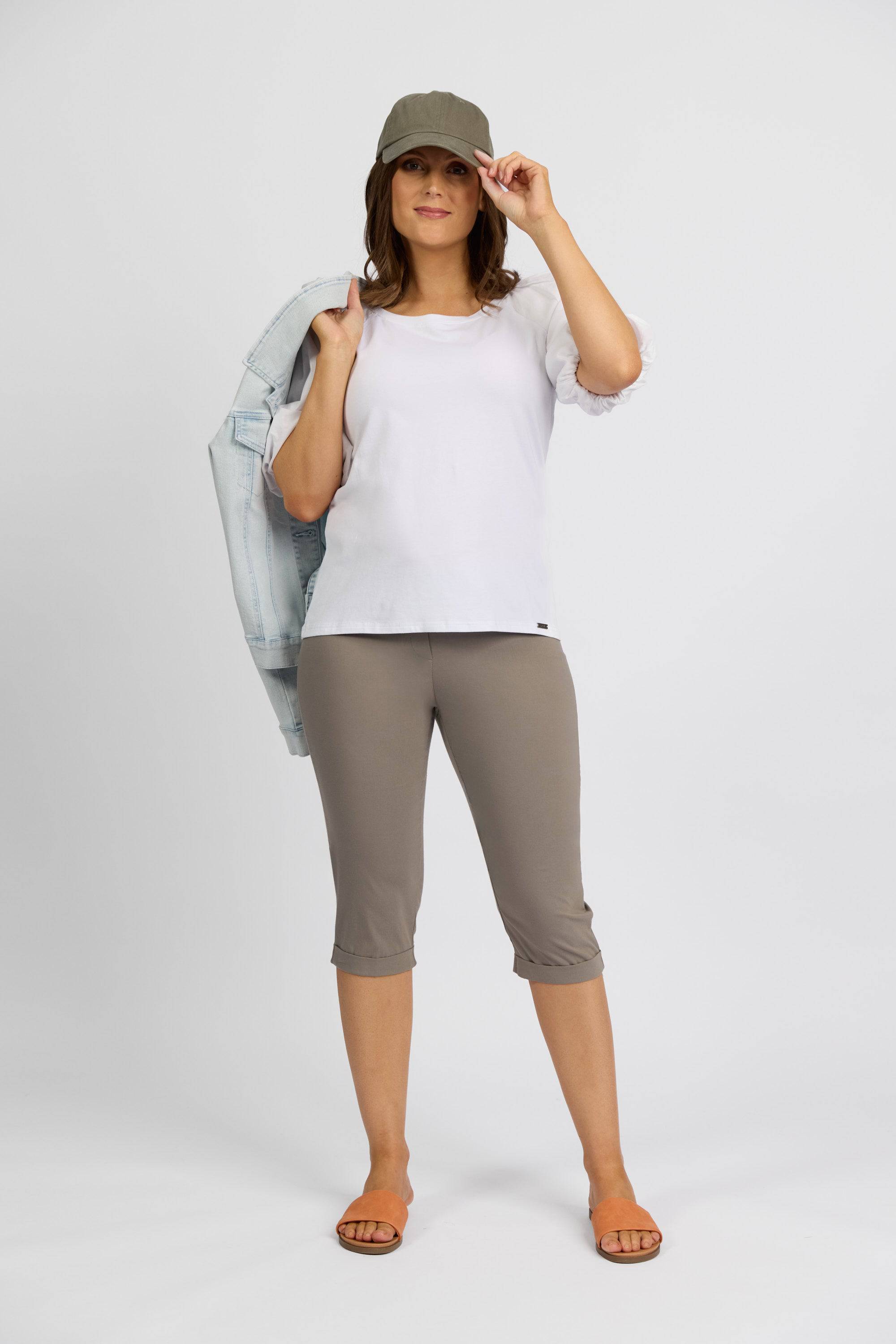 A woman stands against a plain background wearing a white shirt, Vassalli Paris Short in Latte, sandals, a green cap, and holds a light denim jacket over her shoulder while smiling slightly.