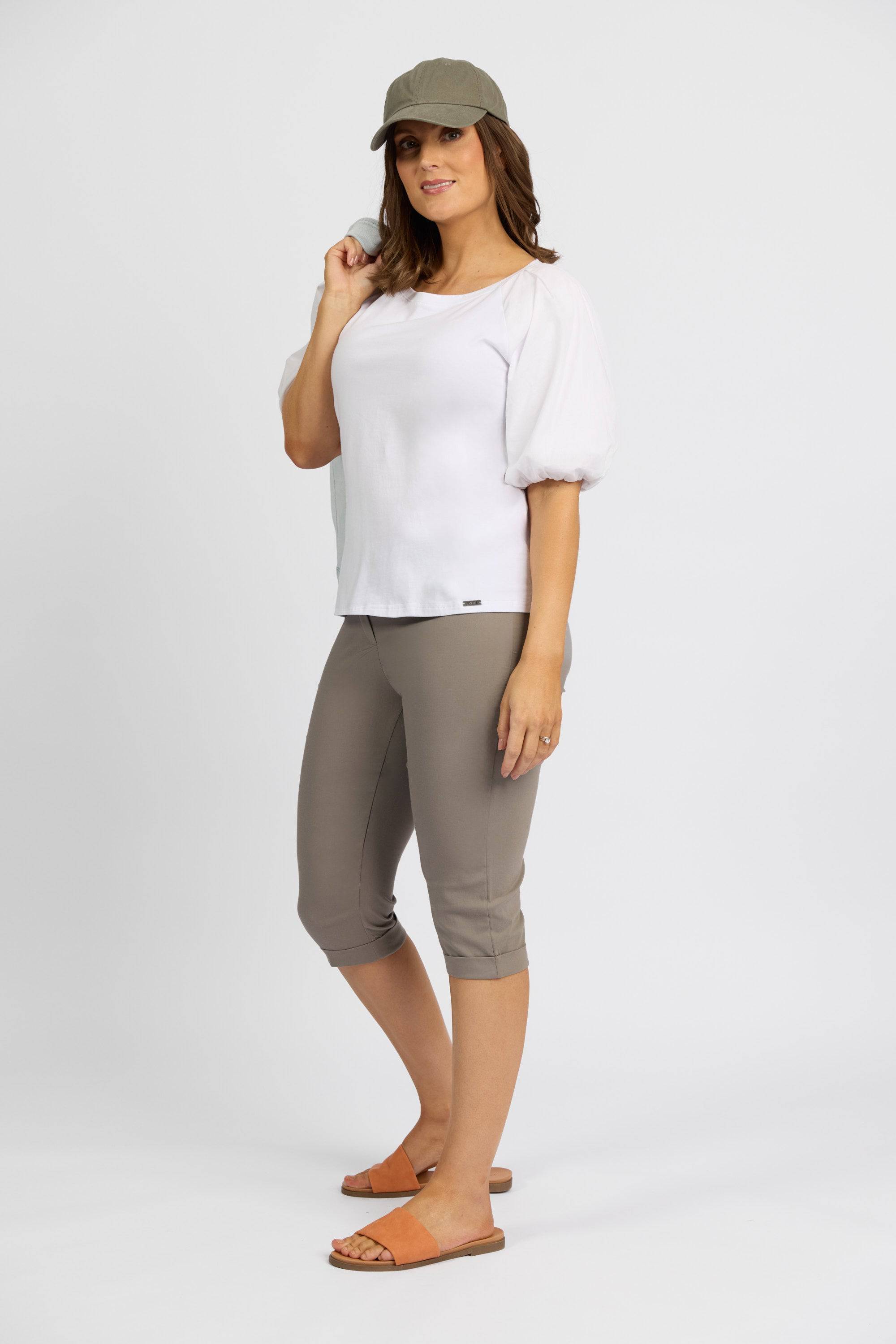 A woman smiles against a white background in a light gray cap, puff-sleeve top, and Vassalli's Paris Short - Latte, paired with tan slide sandals. She holds her cap with one hand.