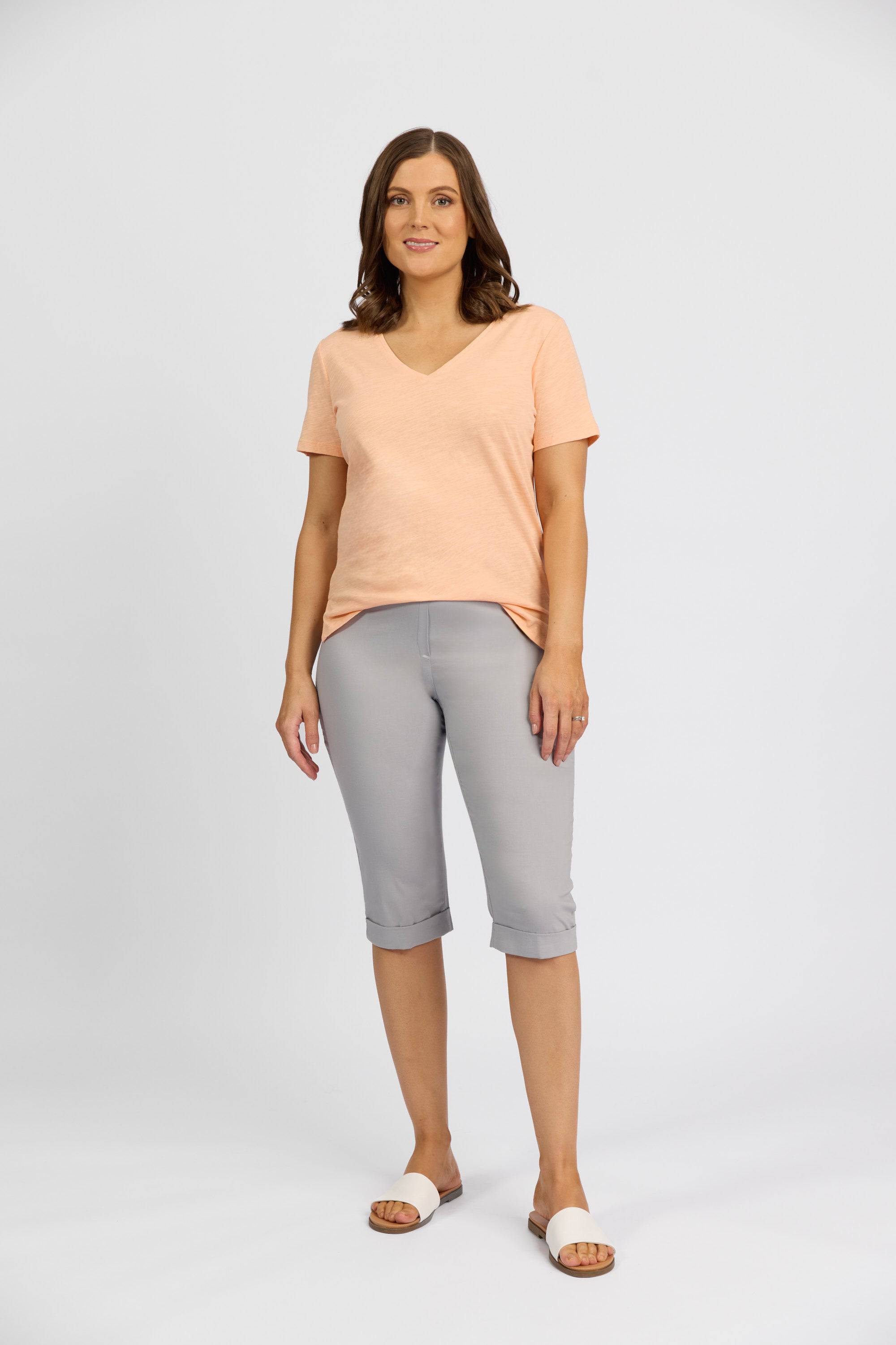 A woman with shoulder-length brown hair and a neutral expression wears Vassalli’s Paris Short in Silver, styled with a peach V-neck t-shirt and white slide sandals against a plain white background.