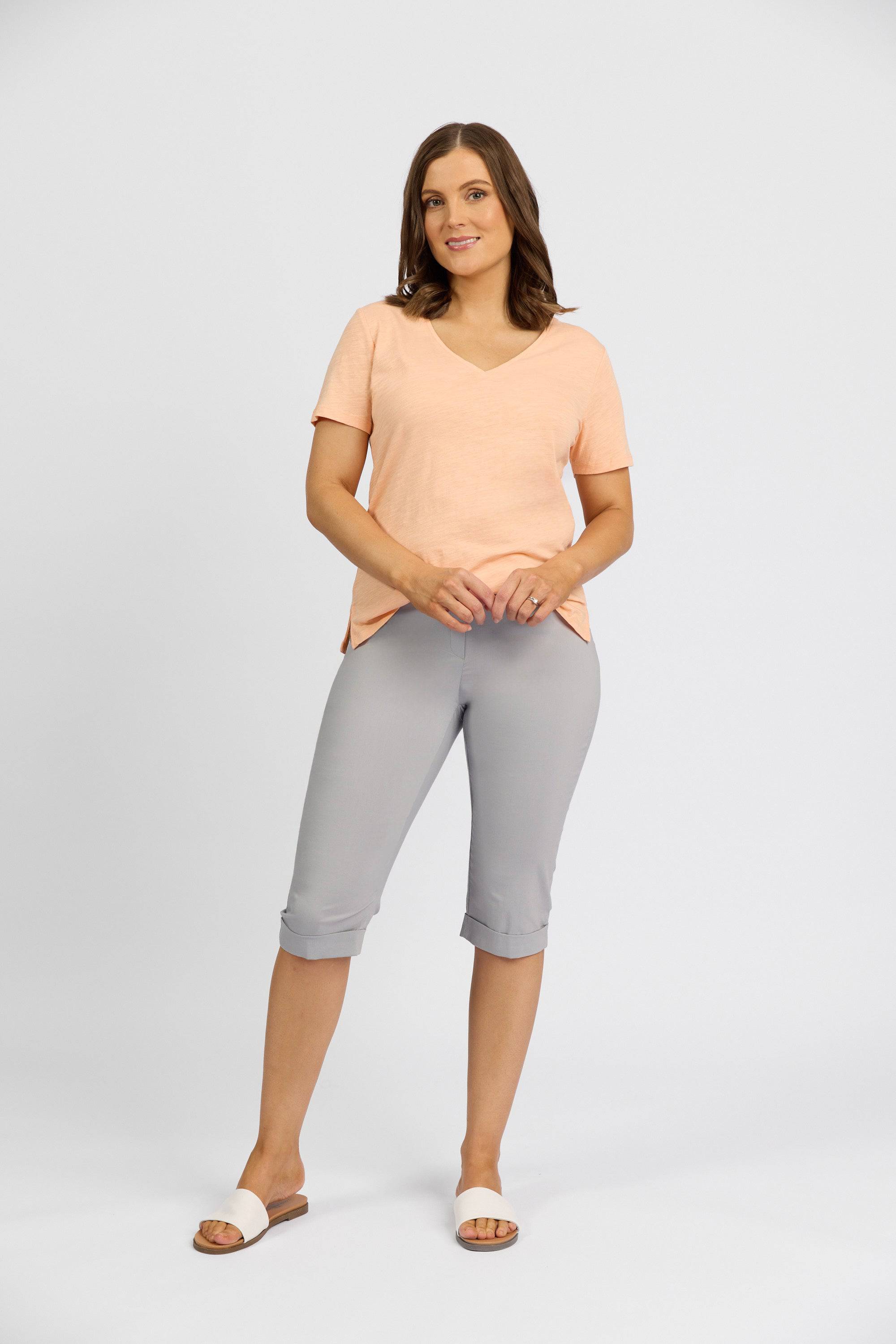 A woman with medium-length brown hair and a slight smile stands against a plain background, wearing the Vassalli Paris Short in Silver and a peach V-neck T-shirt, paired with white slide sandals.