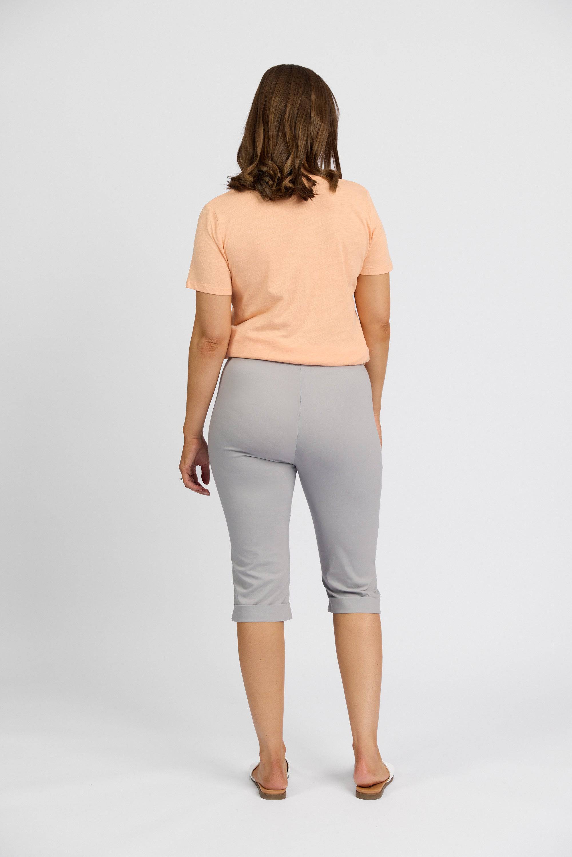 A woman with shoulder-length brown hair stands facing away against a plain white background, wearing a peach short-sleeve shirt, sandals, and Vassalli’s Paris Short - Silver mid-rise stretch capri pants.