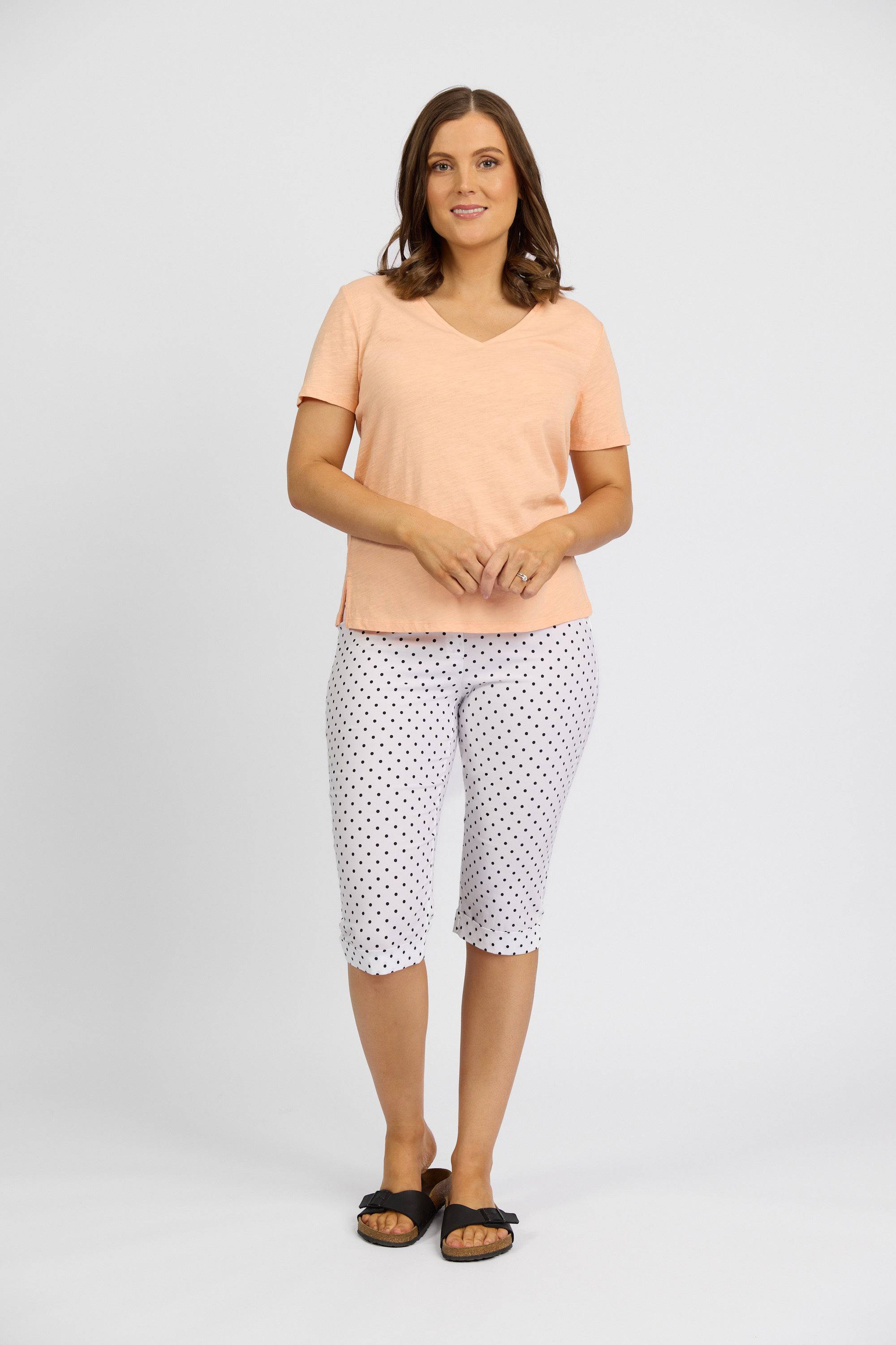 A woman poses against a white background wearing Vassalli Paris Printed Short - Spot, styled with a light peach V-neck top and black slide sandals. She smiles, hands clasped in front of her.