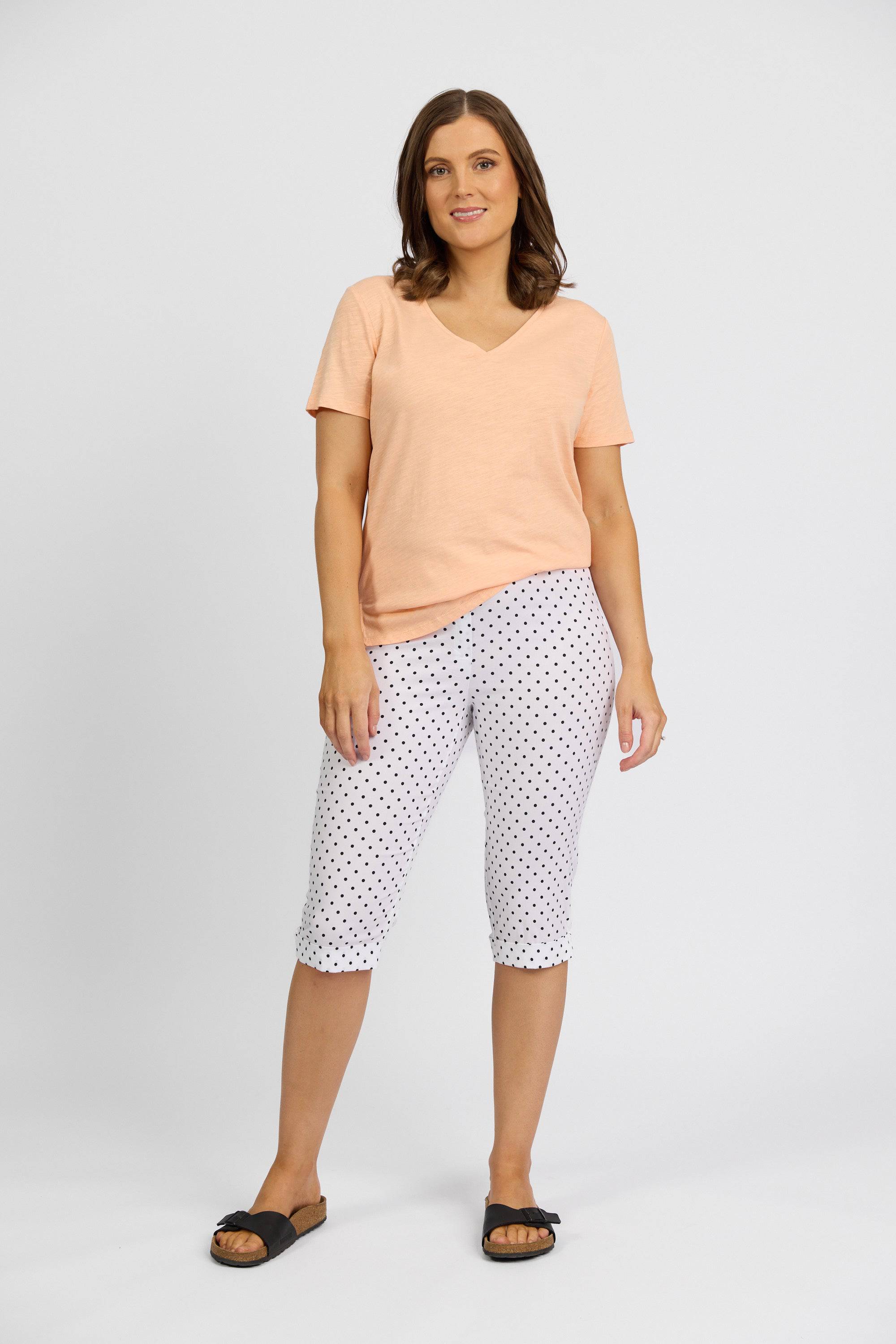 A woman with shoulder-length brown hair and a relaxed expression stands against a plain background, wearing Vassalli's Paris Printed Short - Spot in white with polka dots, a peach short-sleeve V-neck top, and black slide sandals.