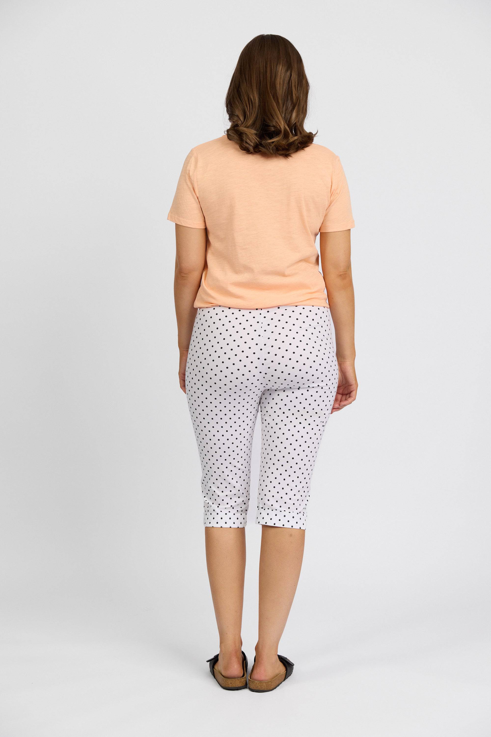 A person with shoulder-length brown hair stands facing away, wearing a peach short-sleeve top, Vassalli Paris Printed Short - Spot capri pants, and black sandals against a plain light background.