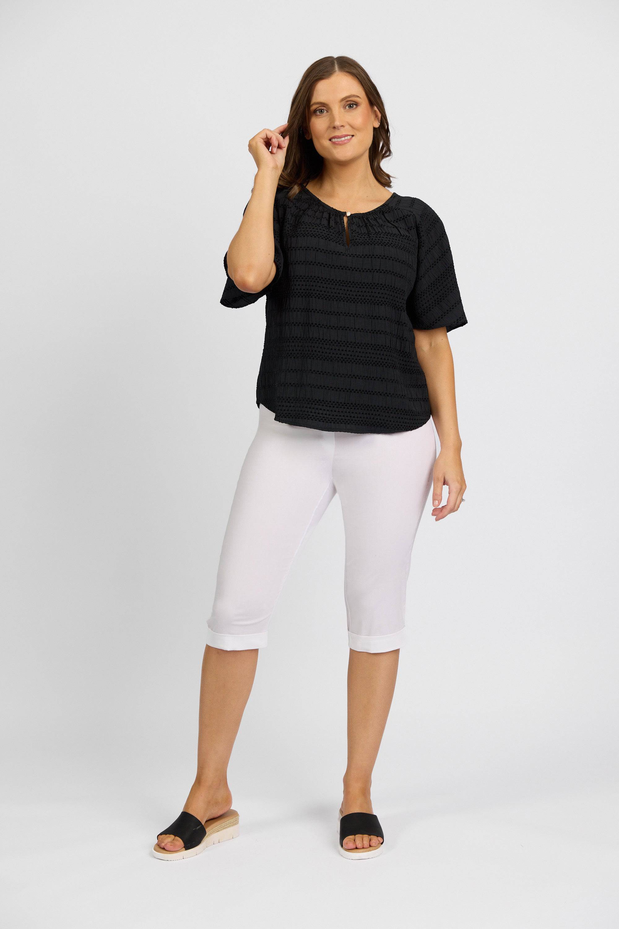 A woman with light brown hair smiles against a plain background, wearing the Vassalli Paris Short - White, a black short-sleeve blouse, and black sandals. She raises one hand near her face.