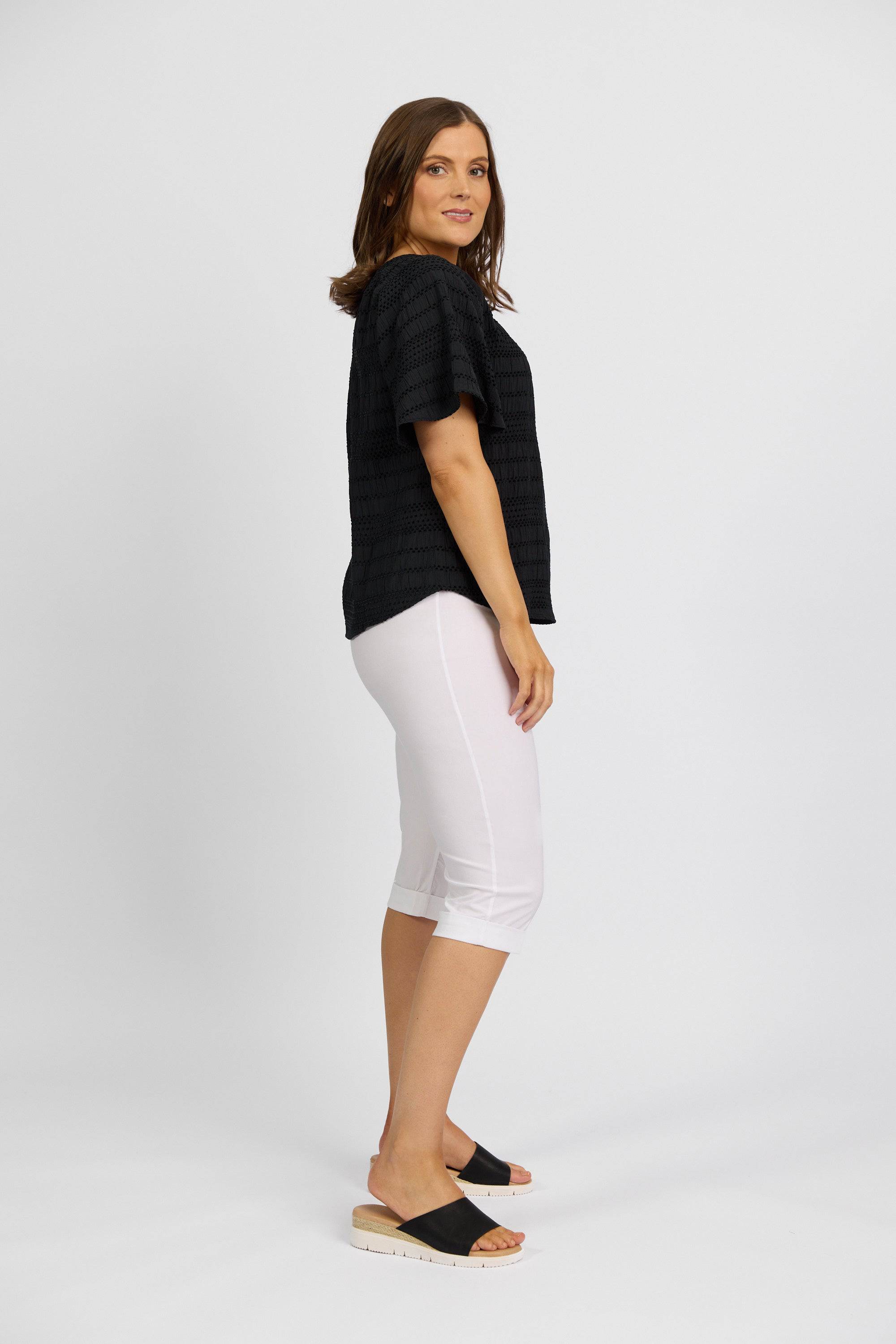 A woman poses sideways against a light background, wearing Vassalli's Paris Short in white, a black stretch short-sleeve top, and black platform sandals.