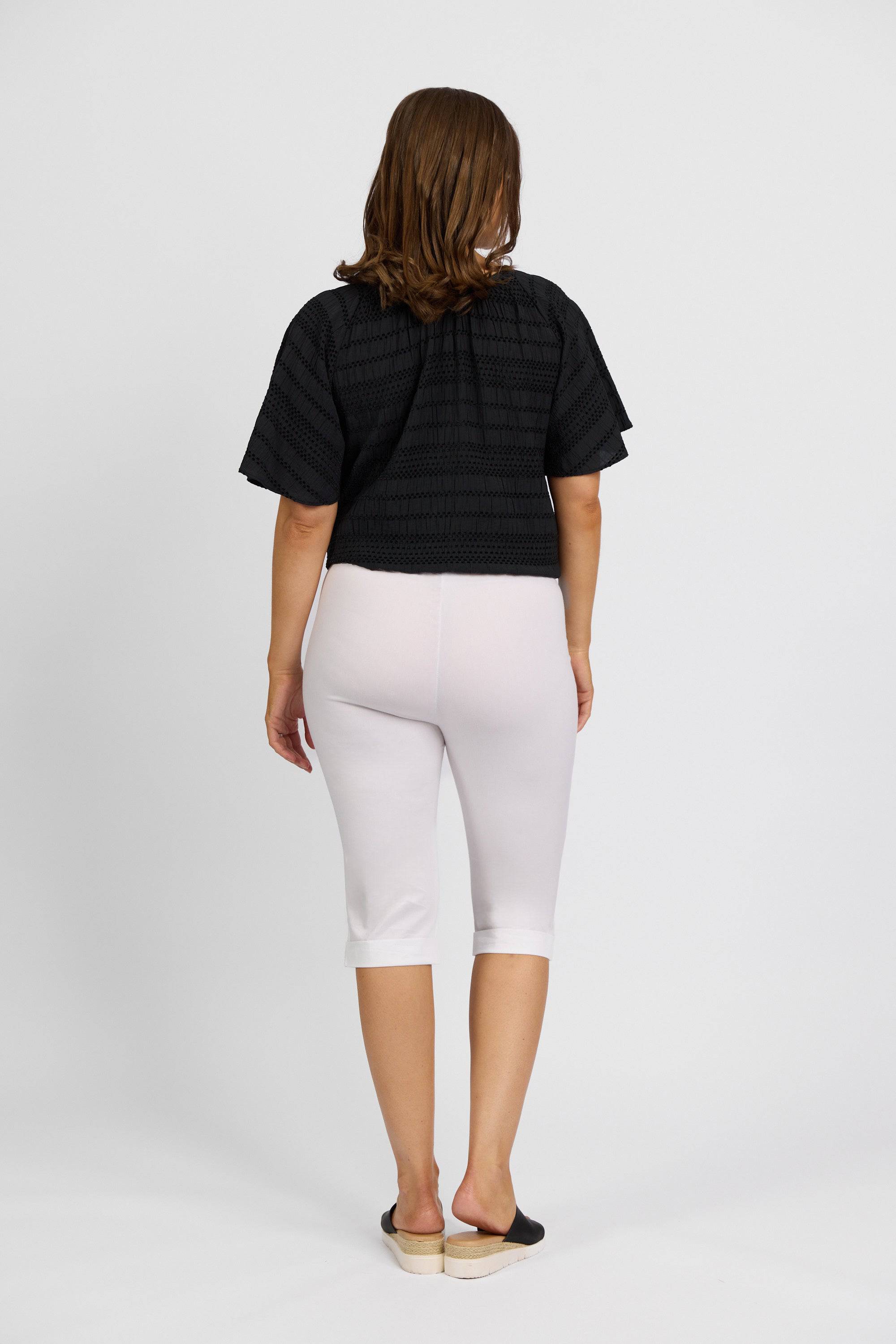 A person with shoulder-length brown hair, wearing a black short-sleeve top and Vassalli Paris Short - White, stands facing away from the camera against a plain white background.