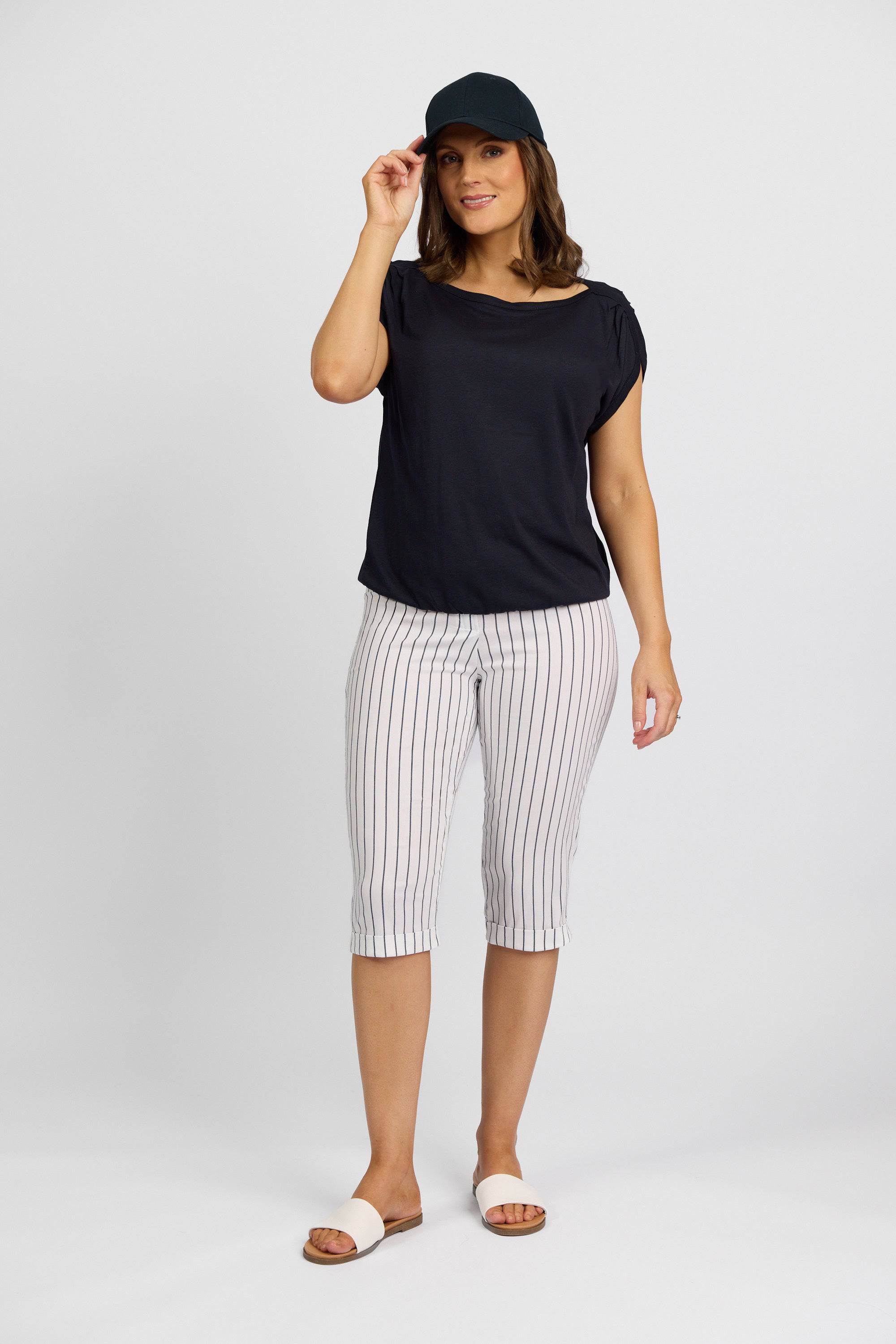 A woman from the Paris Collection wears Vassalli's Paris Printed Short in White w/ Stripe, a black cap, black stretch short-sleeve top, and white slide sandals as she smiles and adjusts her cap against a white background.