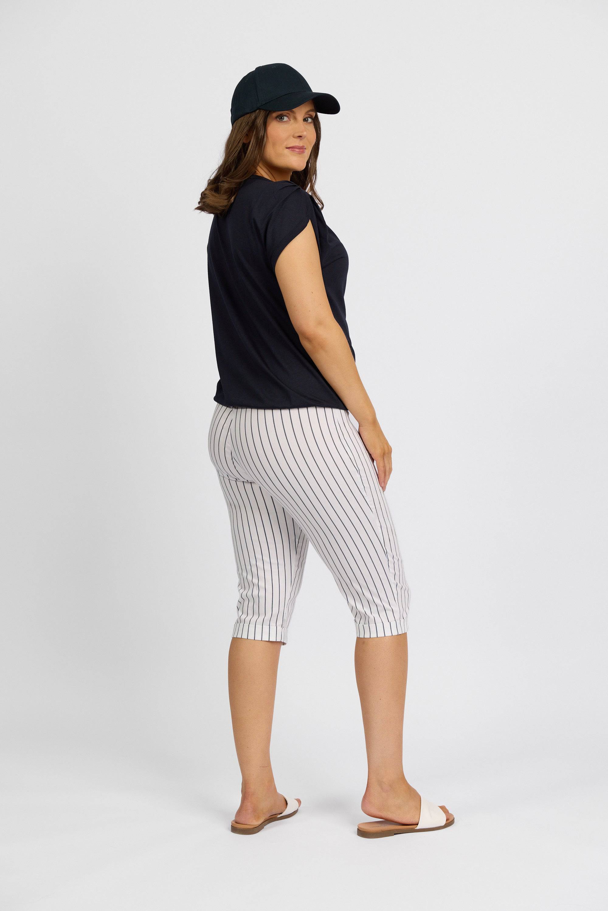 A woman stands turned slightly to the side, wearing a black cap, black t-shirt, Vassalli Paris Printed Short in white with stripes, and white slide sandals against a plain white background.
