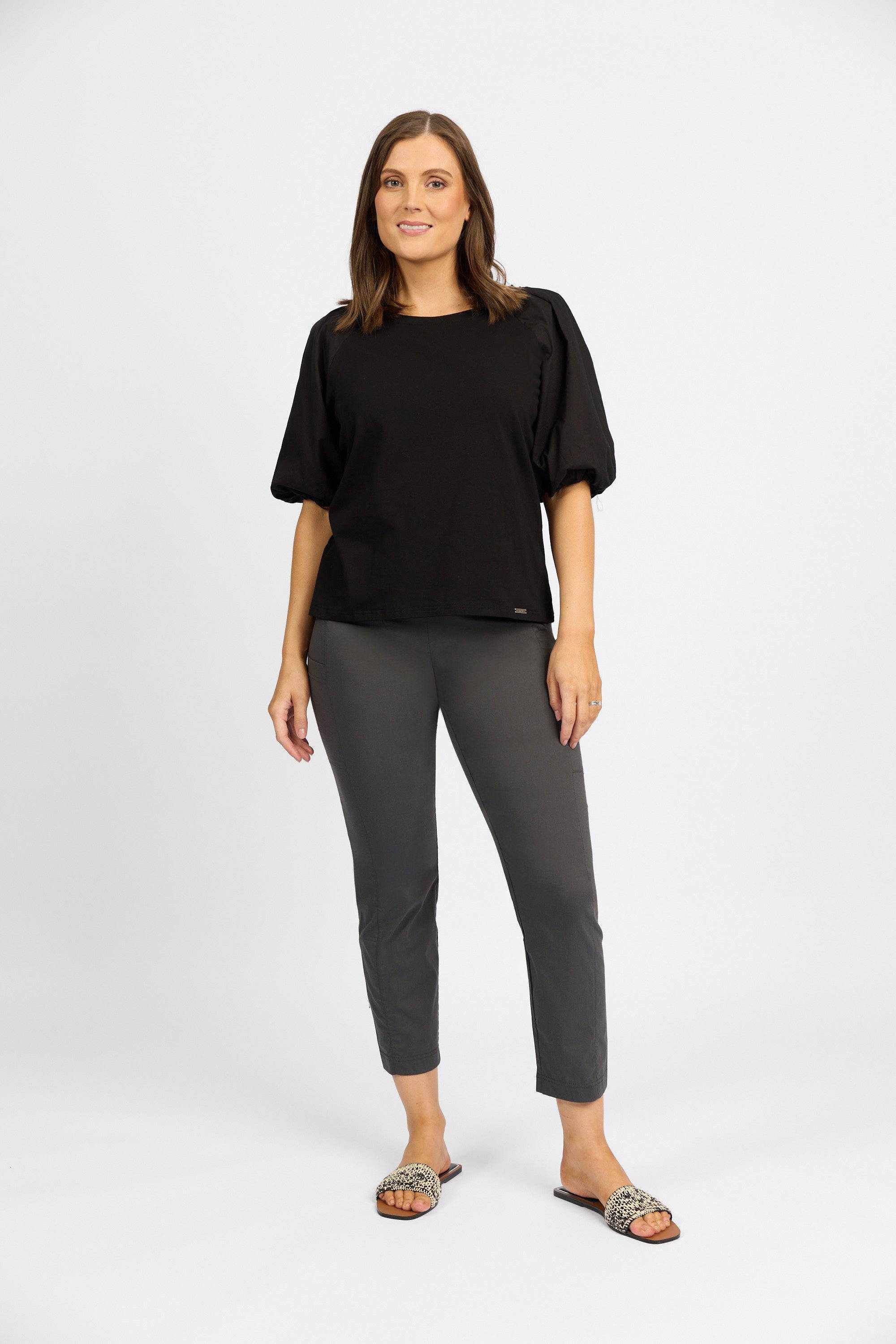 A woman with straight brown hair and a friendly expression stands against a plain light background, wearing Vassalli Paris Cargo Capri in Beluga, a loose black top with elbow-length sleeves, and slip-on flat sandals.
