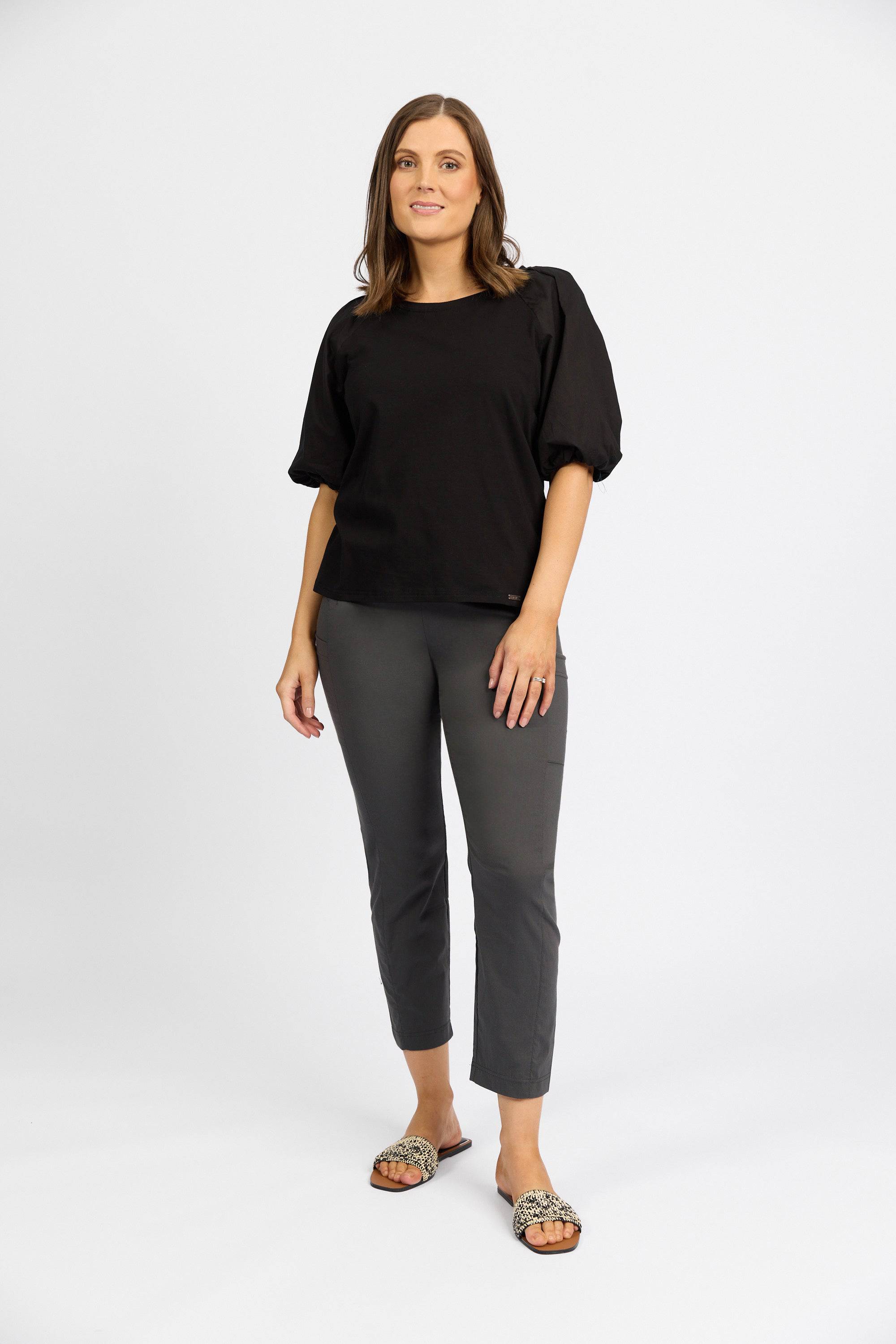 A woman with shoulder-length brown hair faces forward, wearing a loose black top and Vassalli Paris Cargo Capri - Beluga pants, paired with leopard print slide sandals. She poses against a plain white background with a neutral expression.