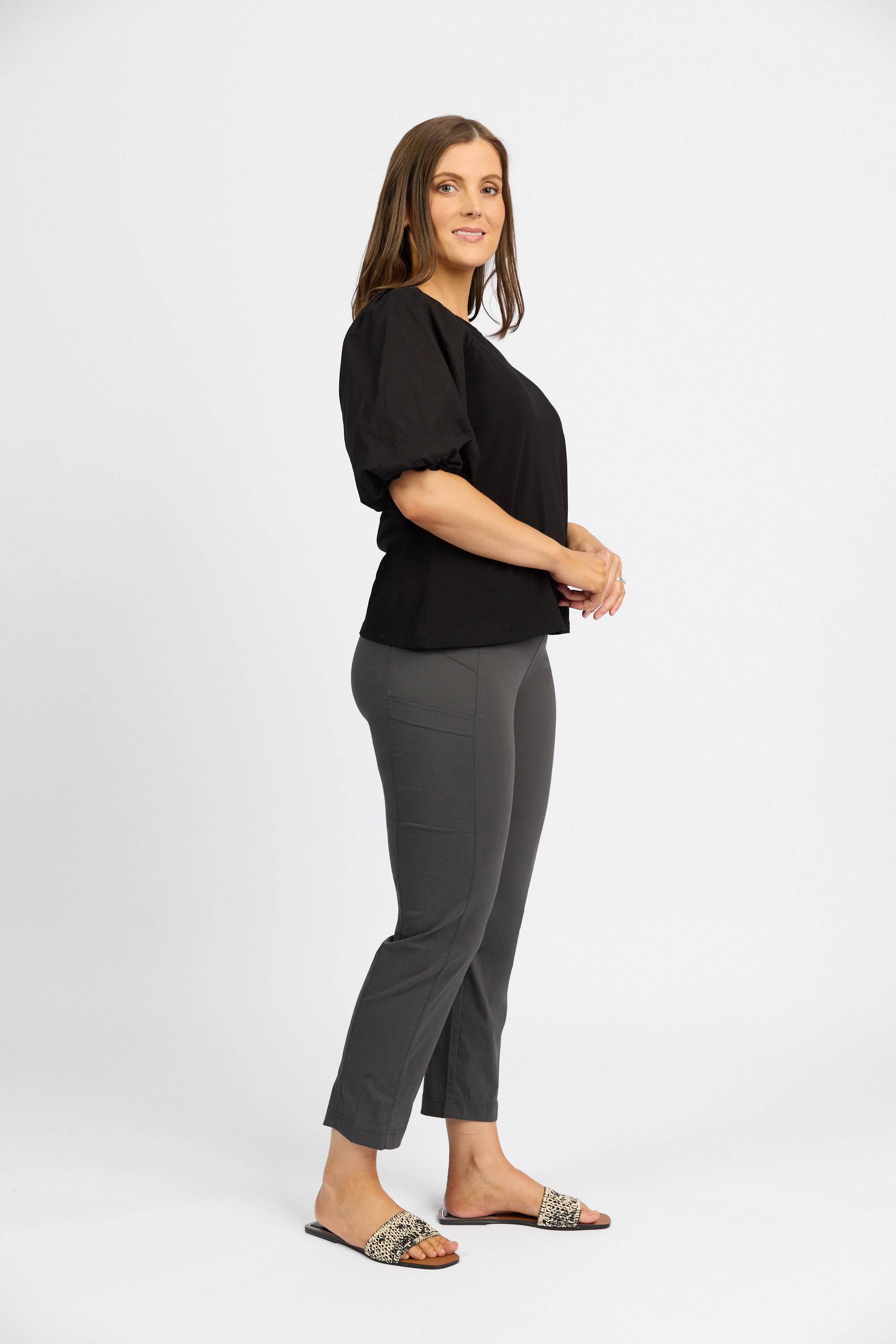 A woman with straight brown hair, wearing a black short-sleeve top and Vassalli Paris Cargo Capri in Beluga, stands sideways in patterned slide sandals, smiling slightly against a plain white background.