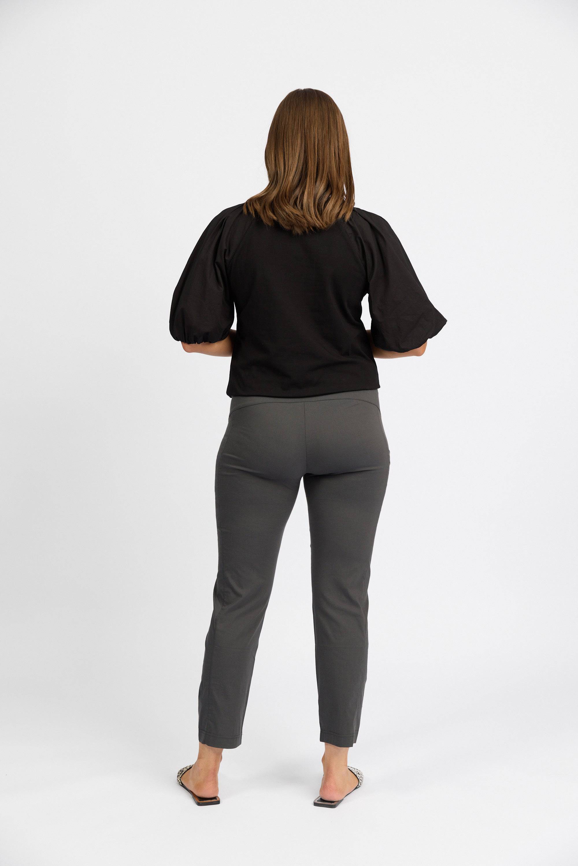 A person with shoulder-length brown hair faces away, wearing a black short-sleeve top, Vassalli Paris Cargo Capri - Beluga pants, and open-toed shoes against a plain white background.