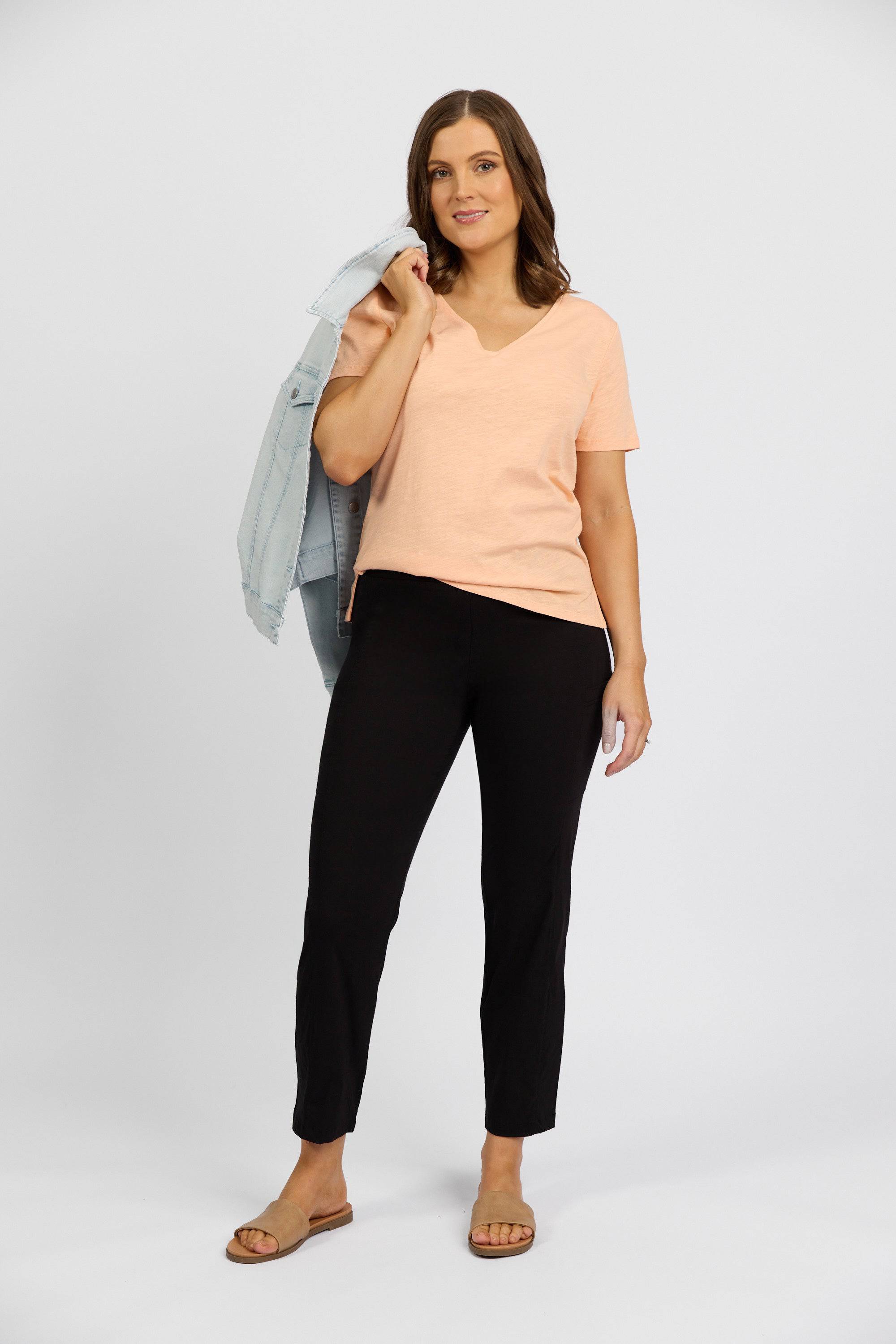 A woman stands against a plain background, smiling at the camera in a peach short-sleeve top, Vassalli Paris Cargo Capri pants in black, sandals, and holding a light blue denim jacket over her shoulder.