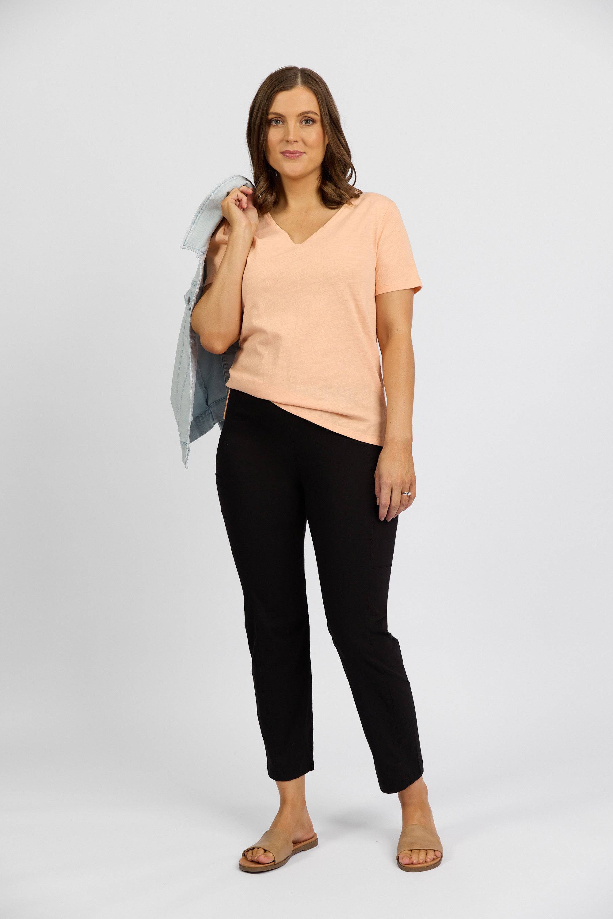 A woman poses against a plain background, holding a light denim jacket. She wears a peach V-neck top, Vassalli Paris Cargo Capri - Black, and brown sandals for a relaxed, casual look.