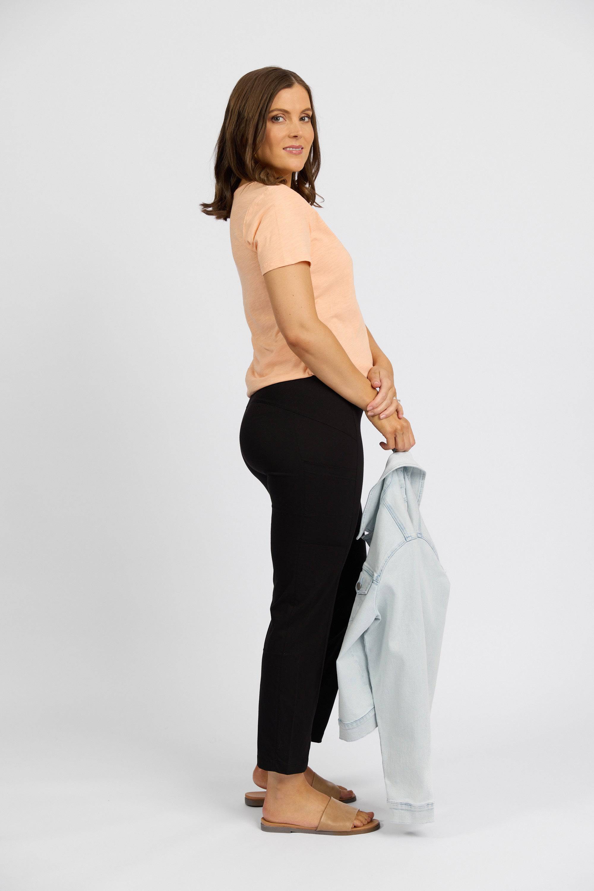 A woman stands sideways against a white backdrop, wearing the Vassalli Paris Cargo Capri - Black with a peach t-shirt and brown sandals. She smiles softly at the camera while holding a light blue denim jacket by her side.