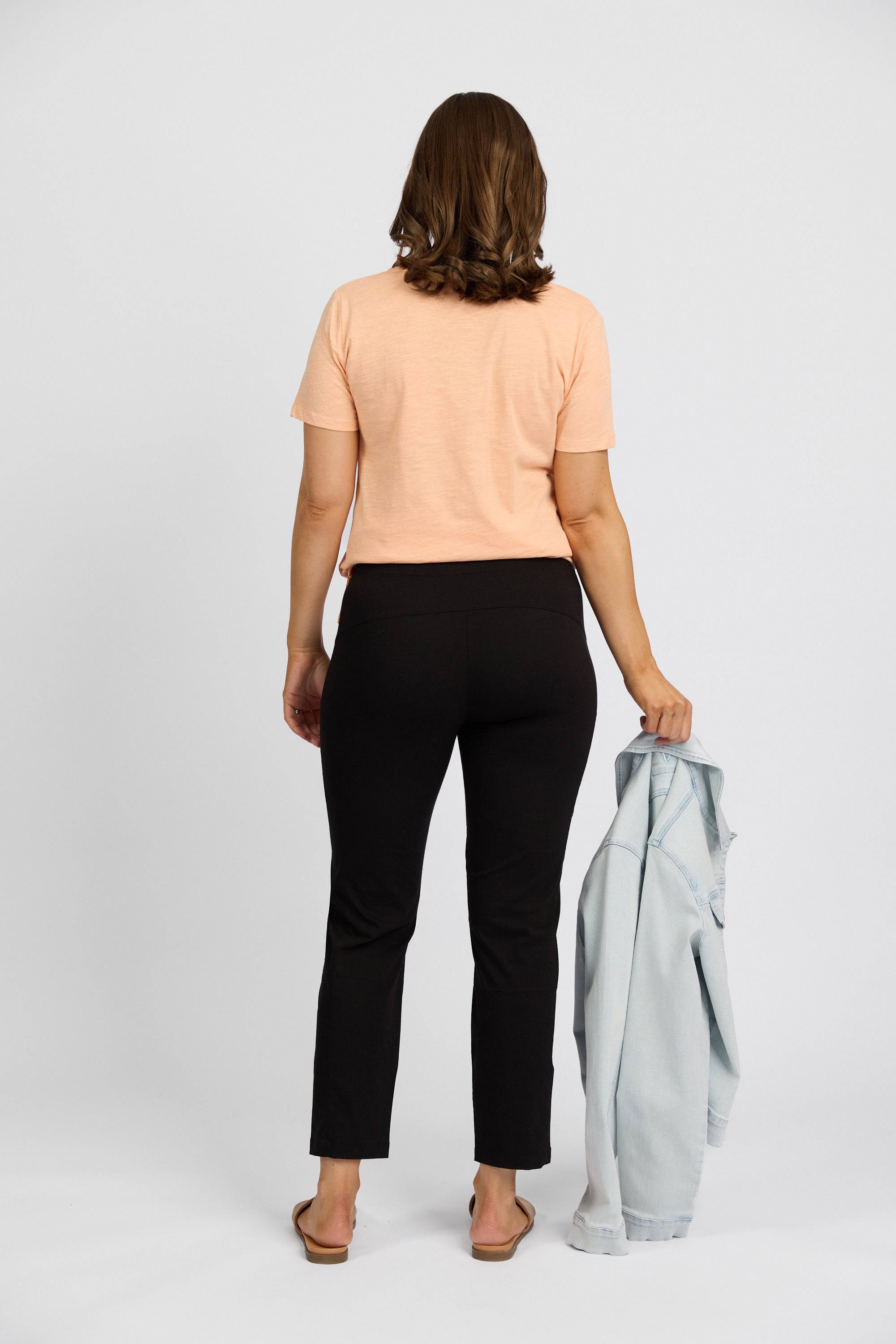 A woman stands facing away, wearing a peach short-sleeve shirt, Vassalli Paris Cargo Capri in black, and sandals, holding a light blue denim jacket in her left hand against a plain white background.