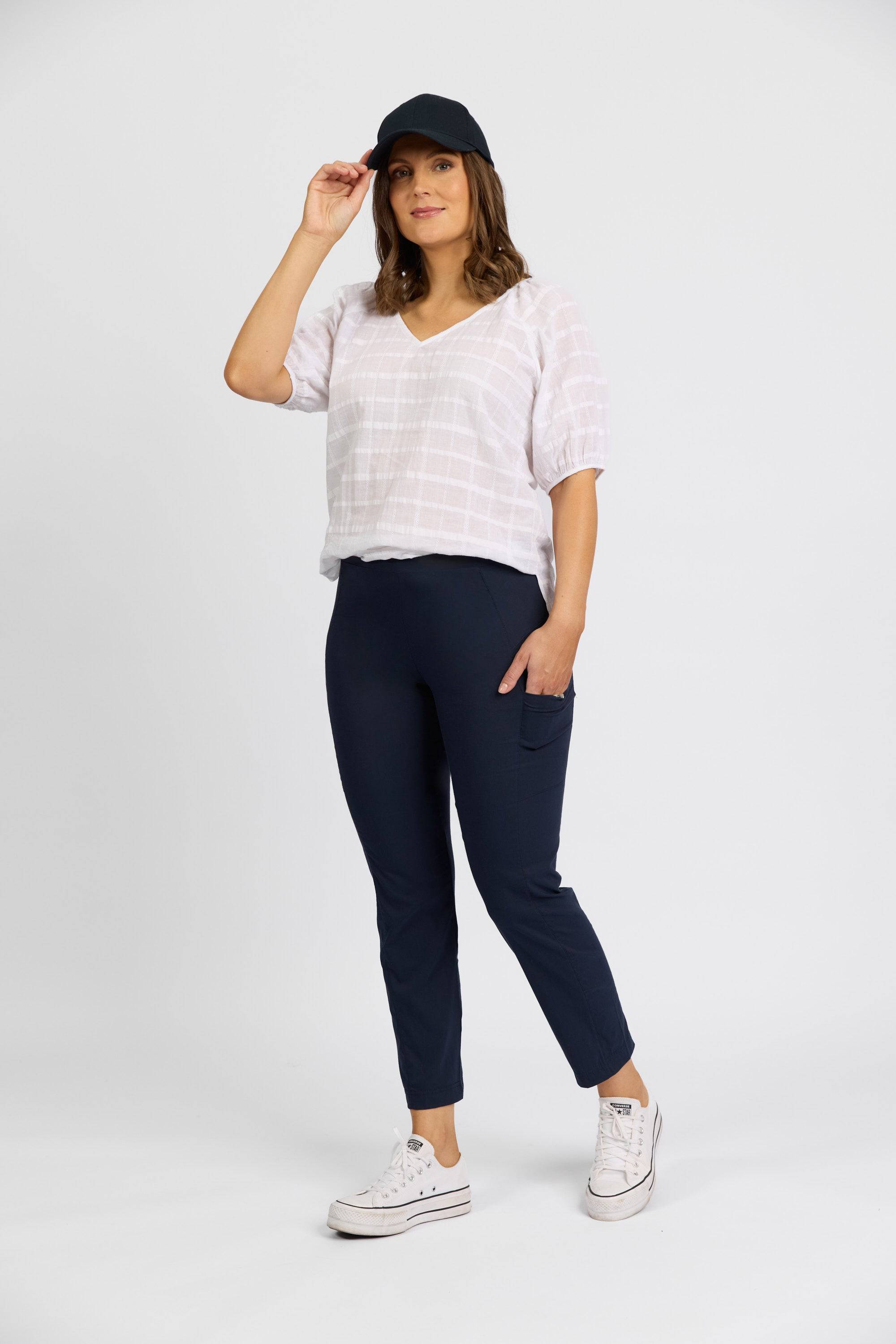 A woman stands against a white background, smiling as she holds her black cap. She wears a white blouse and Vassalli Paris Cargo Capri - Inka pants in navy blue stretch fabric with side pockets—stylish comfort made in New Zealand.