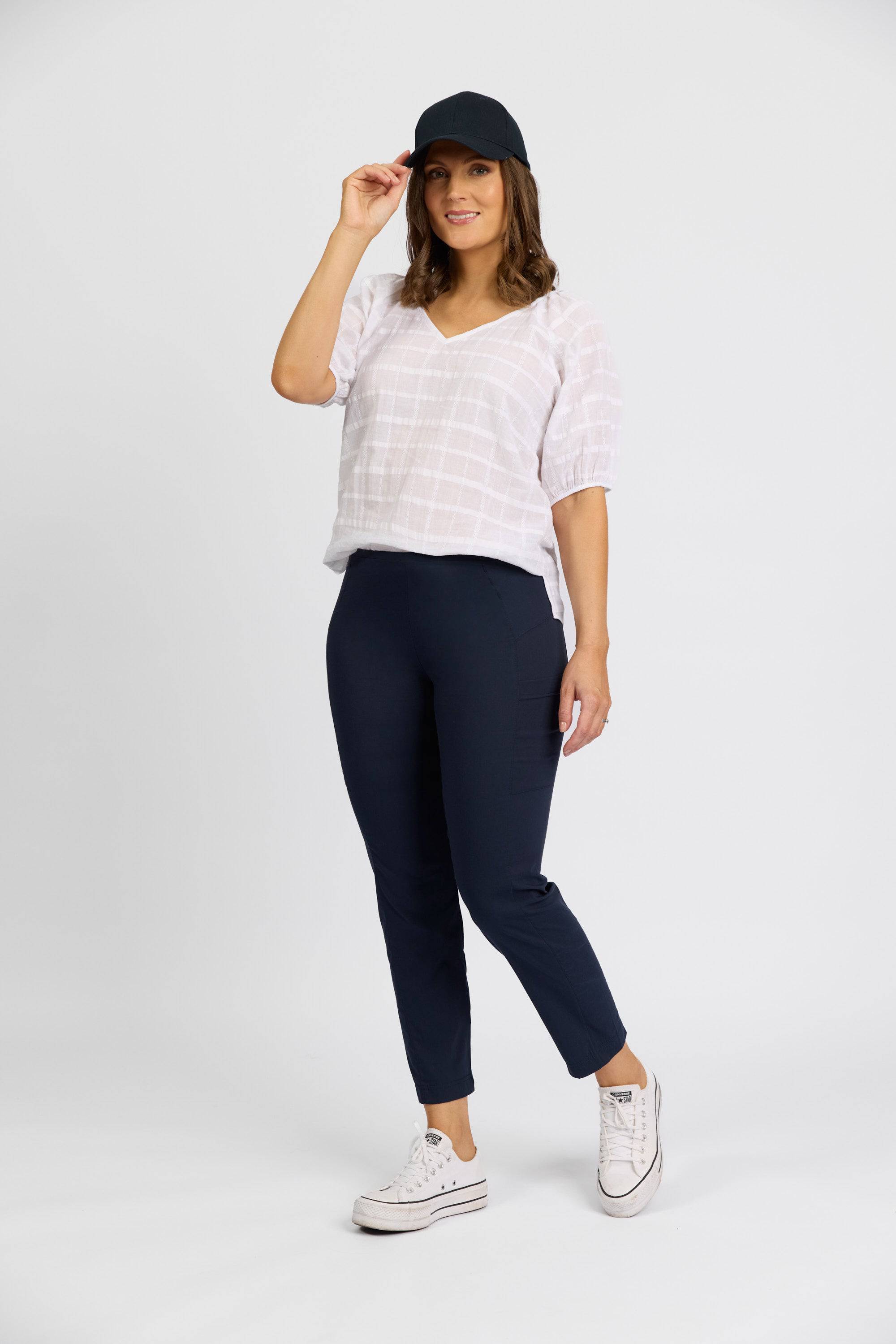 A woman stands against a plain background wearing a white blouse, Vassalli Paris Cargo Capri - Inka pants, white sneakers, and a black cap. She touches the brim of her cap and smiles slightly.
