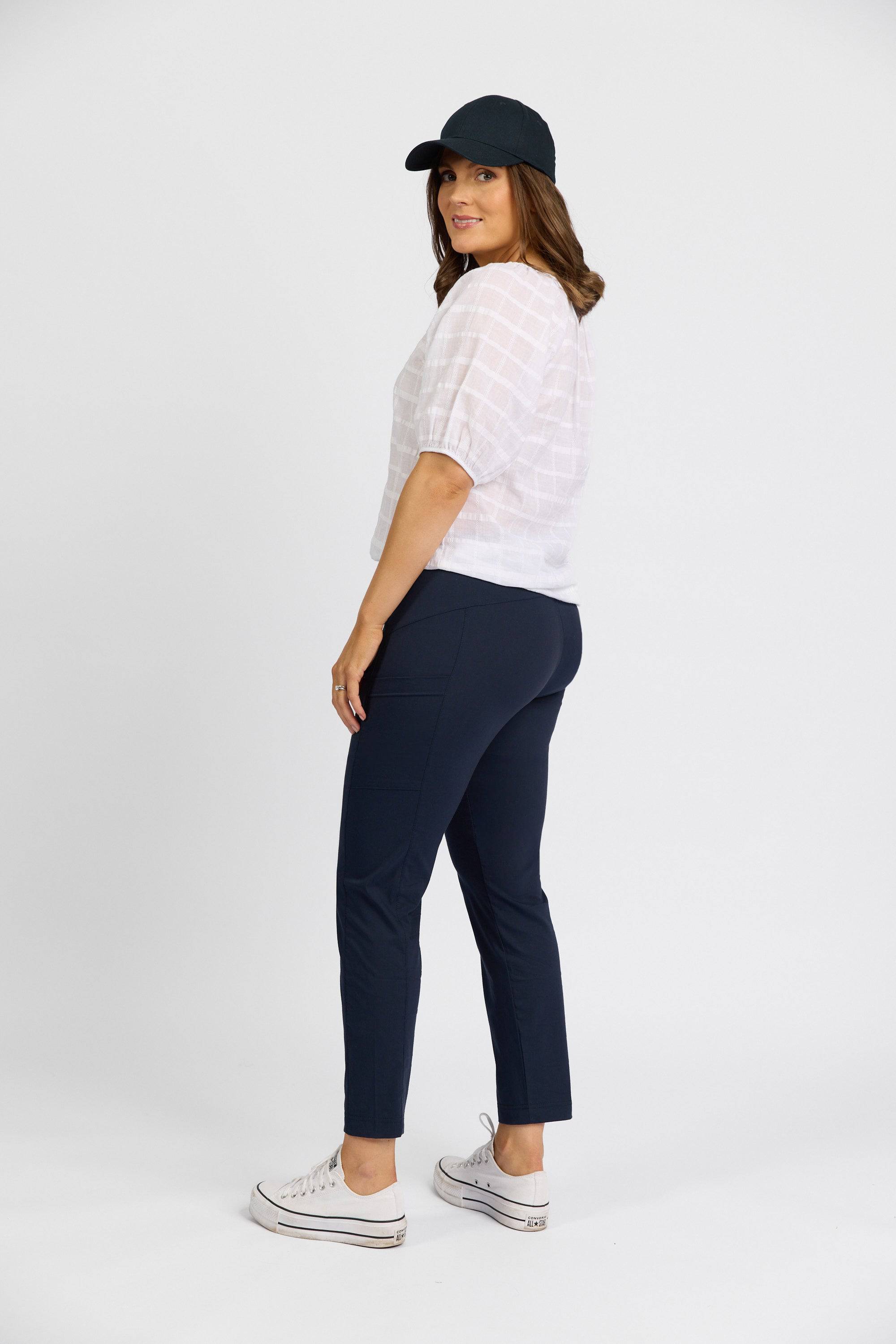 A woman in a black cap, white short-sleeve blouse, Vassalli Paris Cargo Capri - Inka, and white sneakers stands sideways, looking over her shoulder against a plain white background.