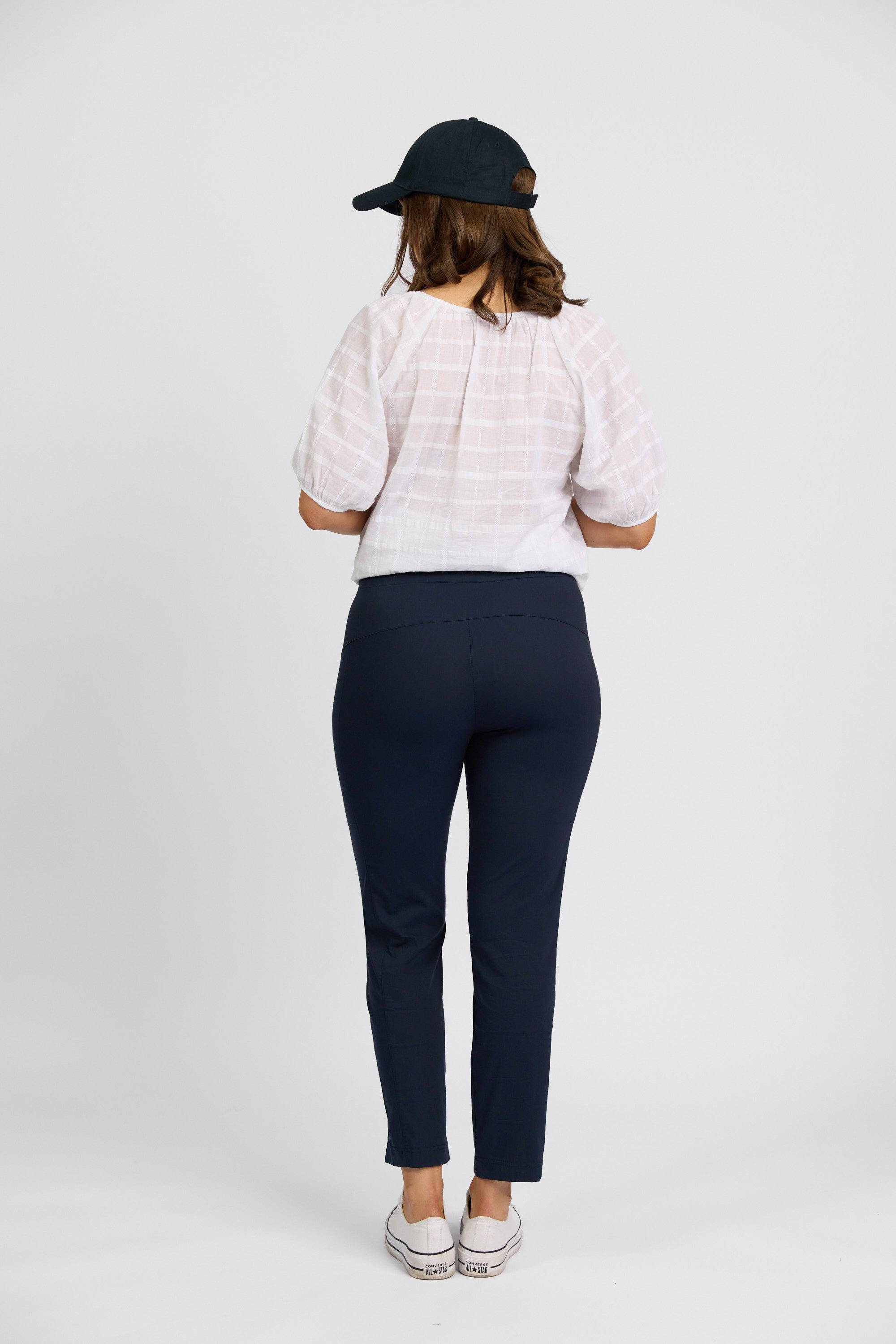 A woman faces away, styled in a black cap, white short-sleeve blouse, Vassalli Paris Cargo Capri - Inka in dark blue stretch fabric, and white sneakers against a plain white background.