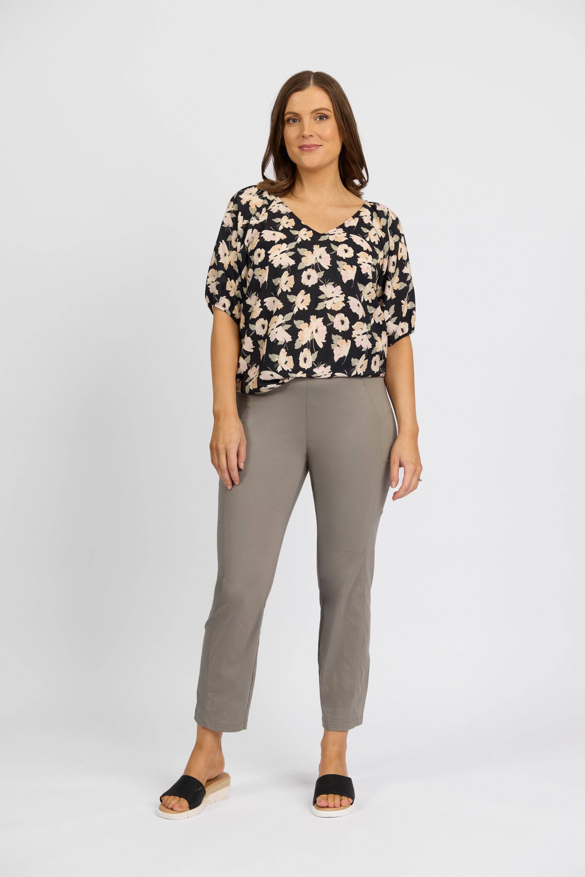 A woman with shoulder-length brown hair and a relaxed expression stands against a plain white background, wearing a Vassalli black floral blouse, Vassalli Paris Cargo Capri pants in latte, and black open-toe sandals.