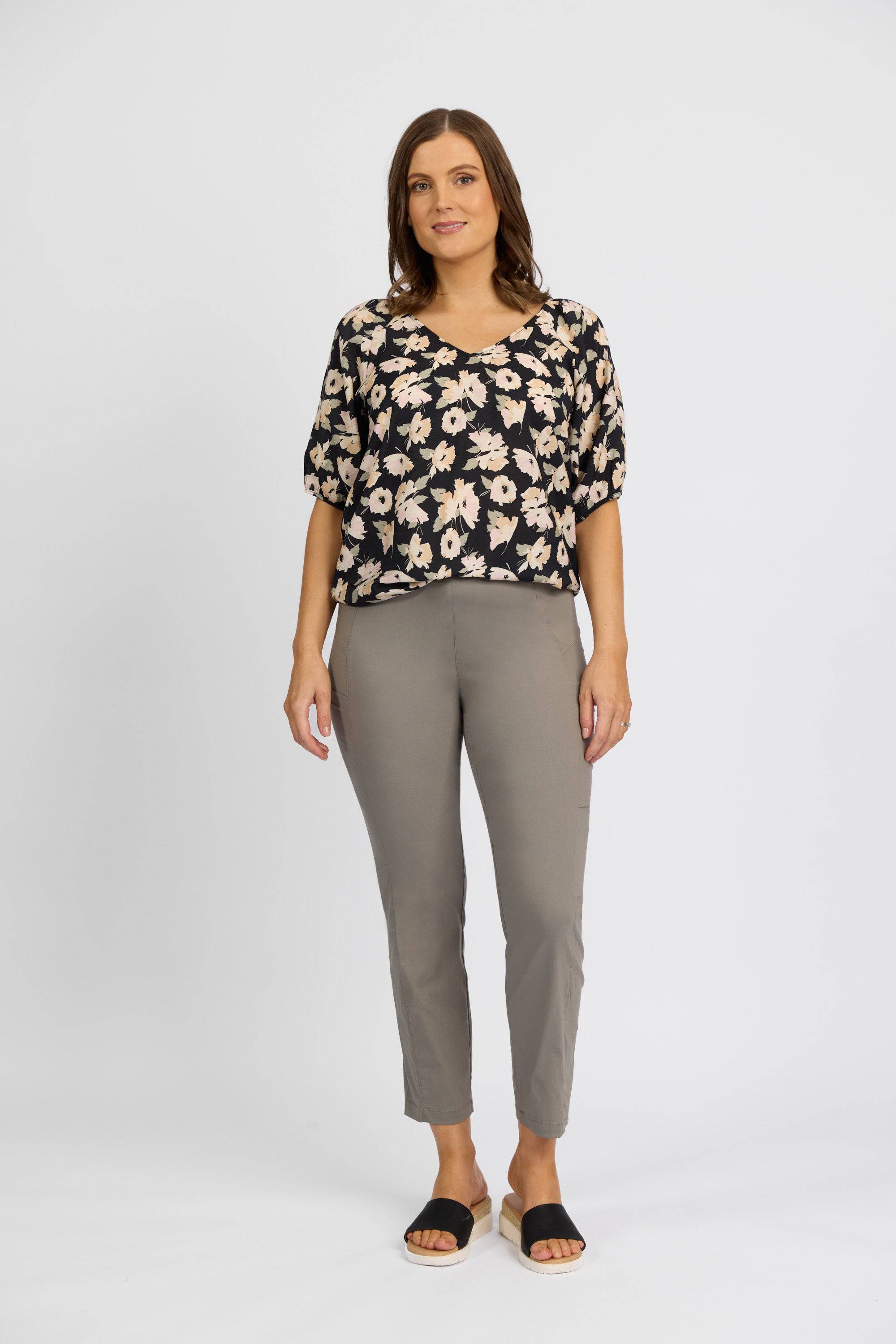 A woman with wavy brown hair and a relaxed expression stands against a plain white background wearing a black floral blouse, Vassalli Paris Cargo Capri pants in Latte, made from stretch fabric, and black slide sandals.
