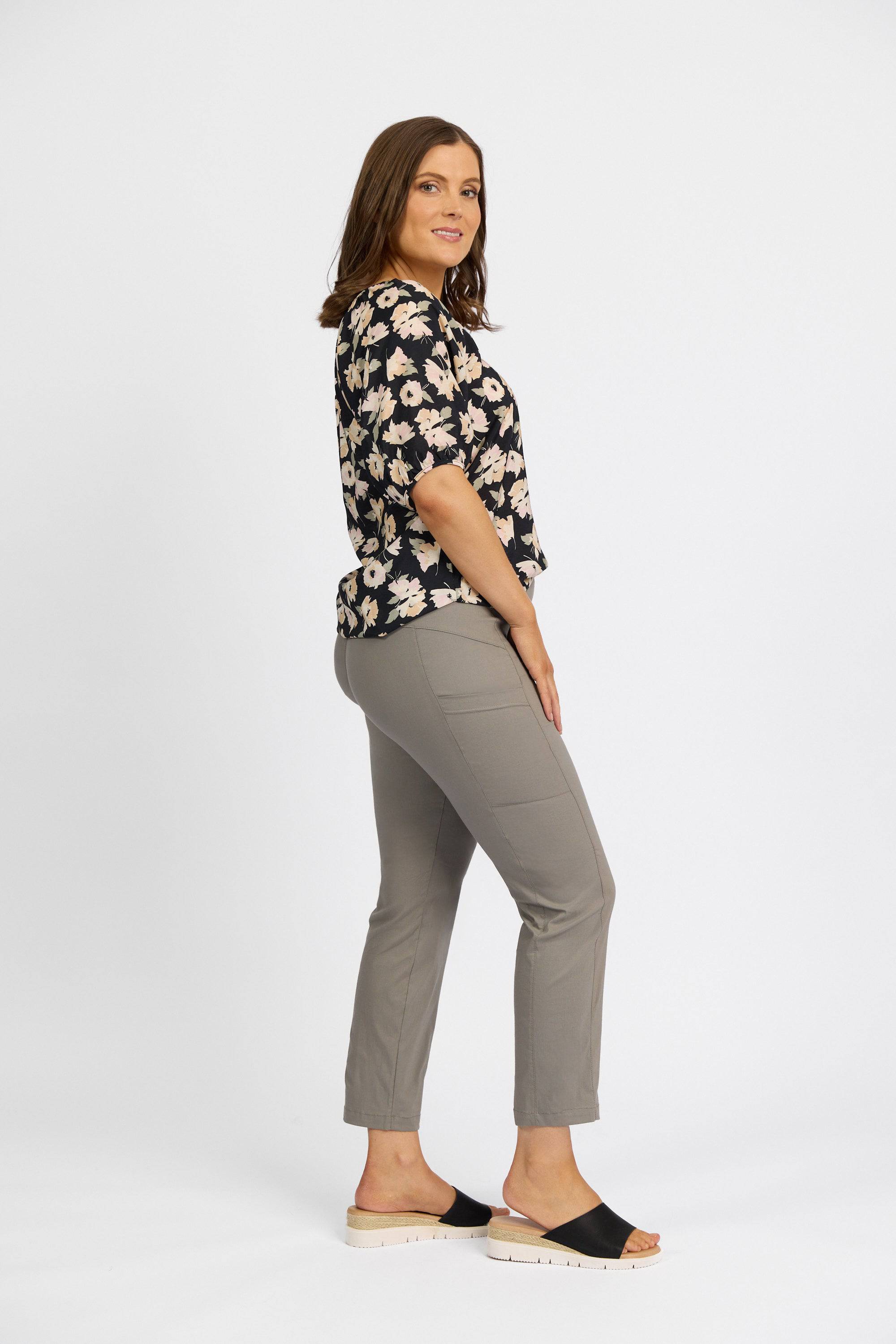 A woman with shoulder-length brown hair stands sideways against a white background, wearing a black floral blouse, Vassalli Paris Cargo Capri pants in Latte, and black slide sandals with white soles, smiling slightly.